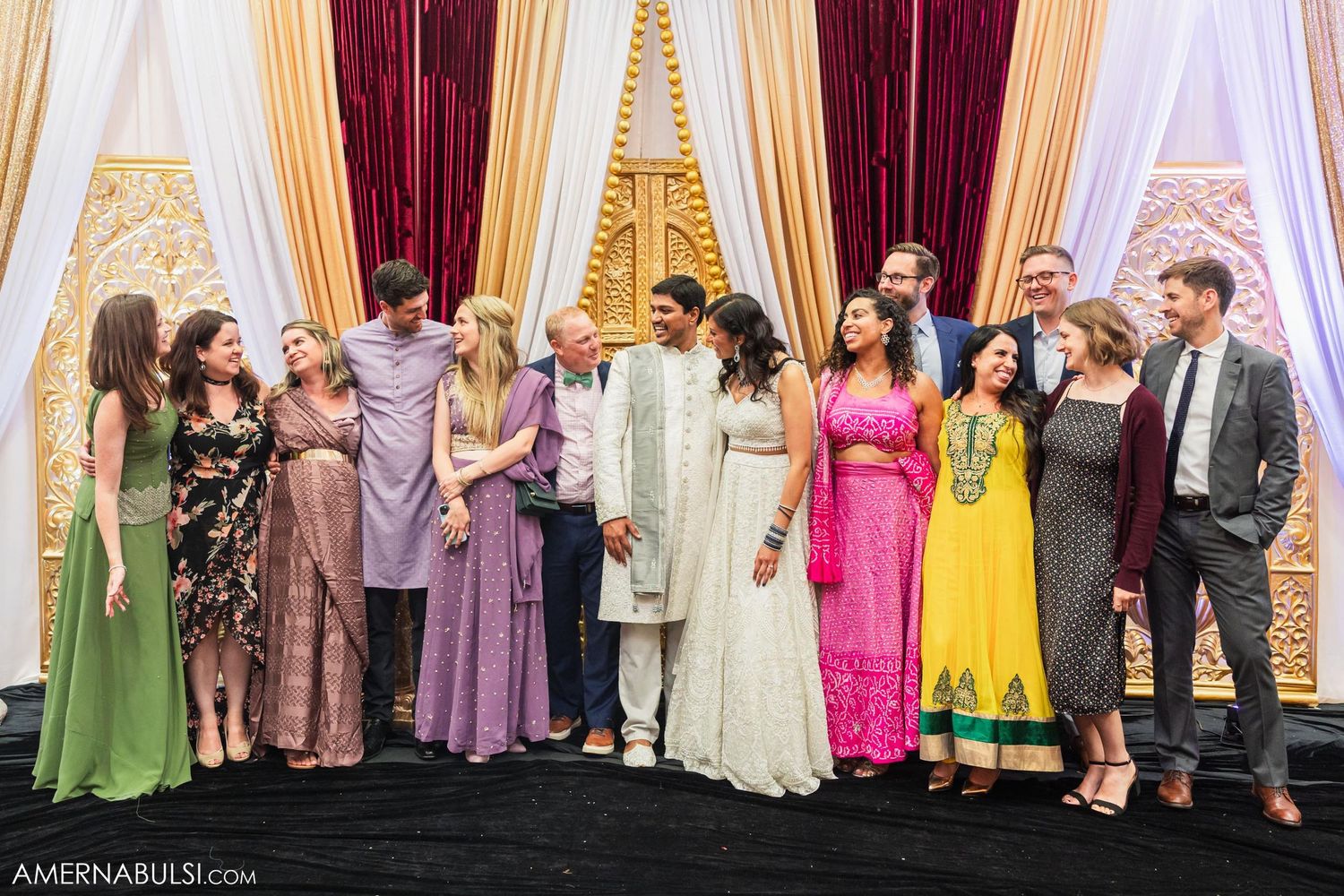 Wedding party members and guests pose together against an elegant backdrop with gold and burgundy drapes.