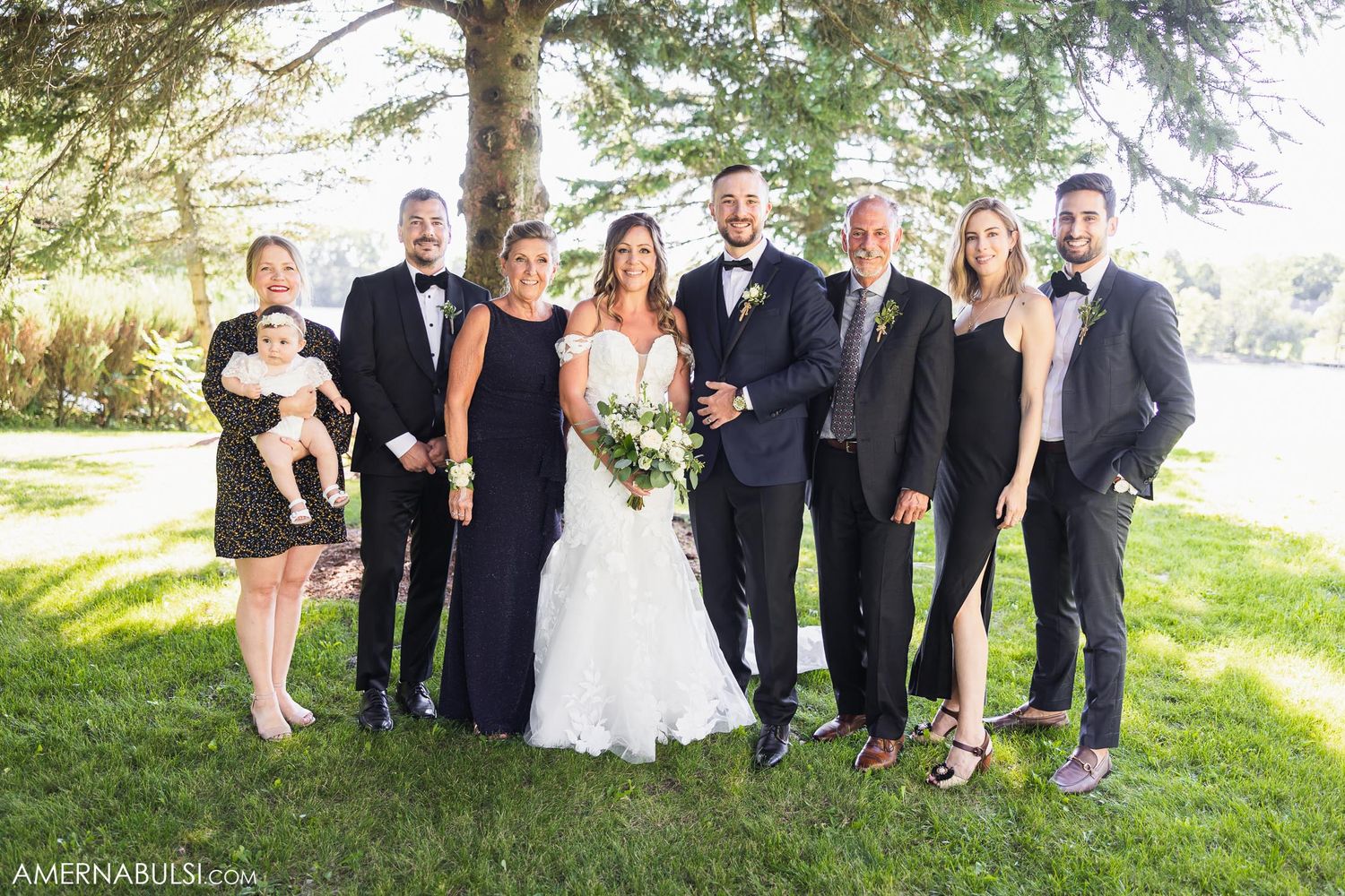 Wedding party poses together on grassy lawn with water view in background.