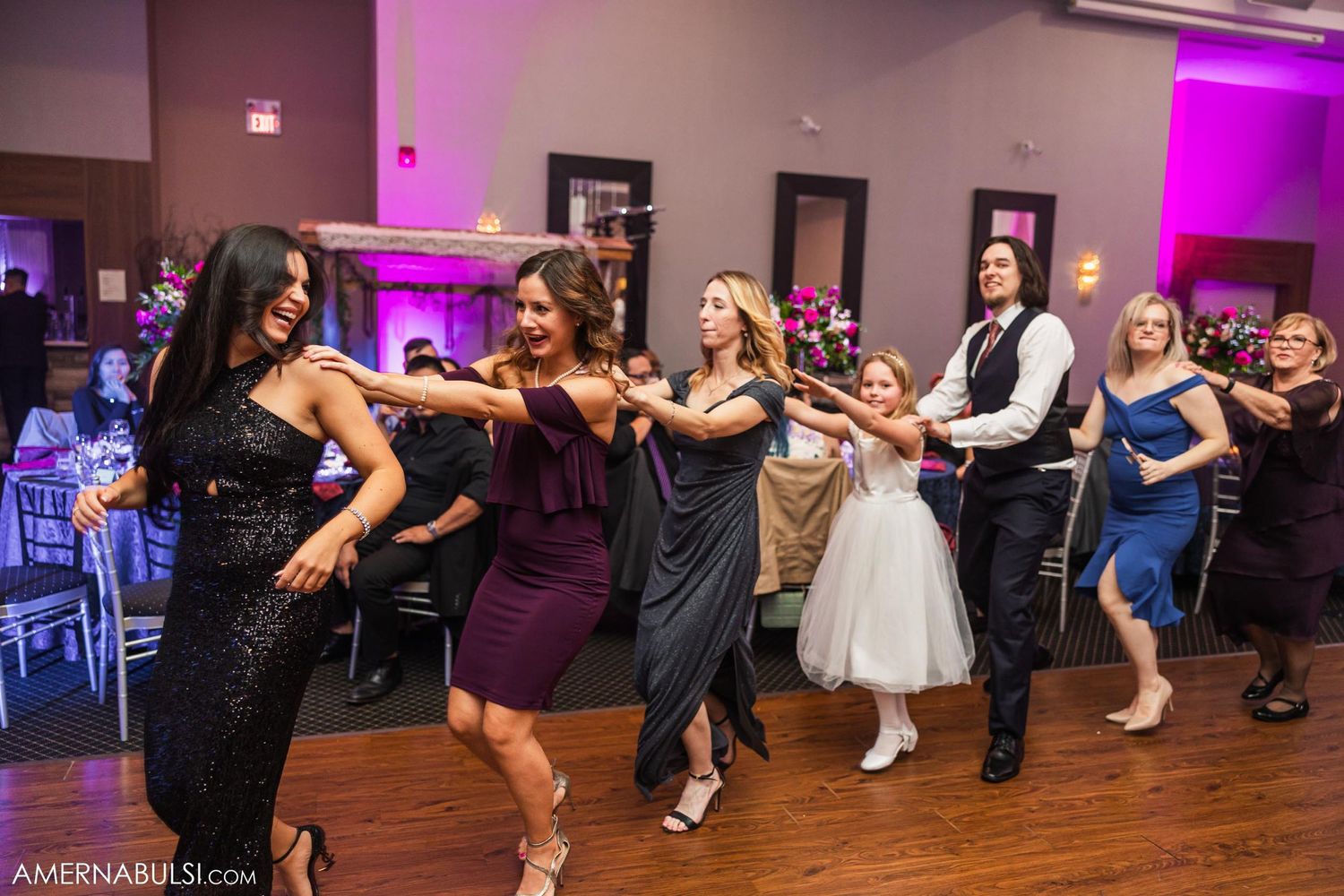 Group of wedding guests performing synchronized dance moves at reception with purple ambient lighting.