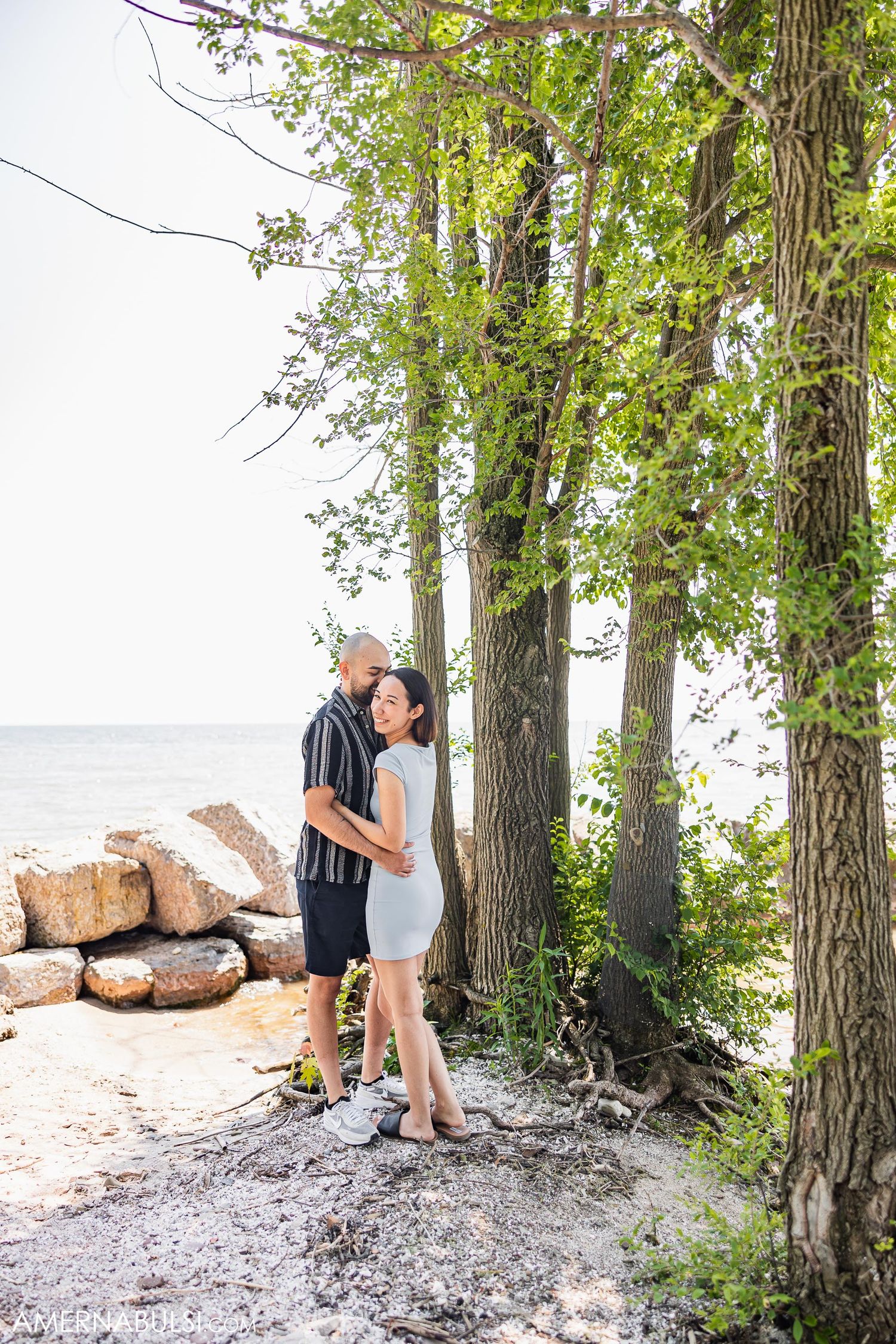 A couple embraces near tall trees along a rocky shoreline.