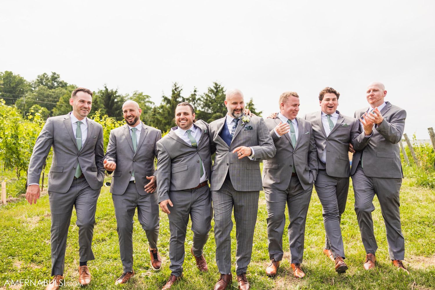 Groomsmen in grey suits jumping and posing together in a vineyard setting.