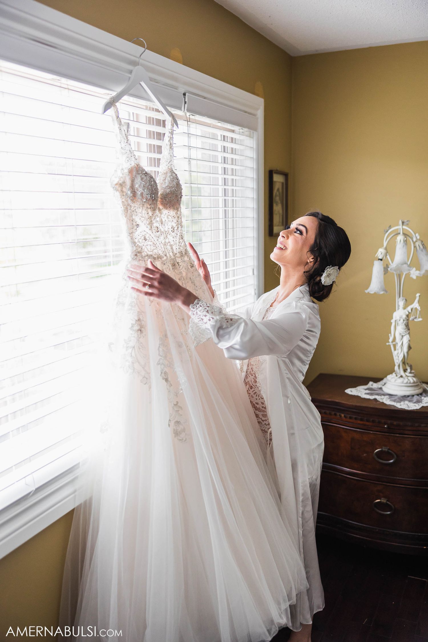 Bride in white robe admiring her wedding dress hanging by window.