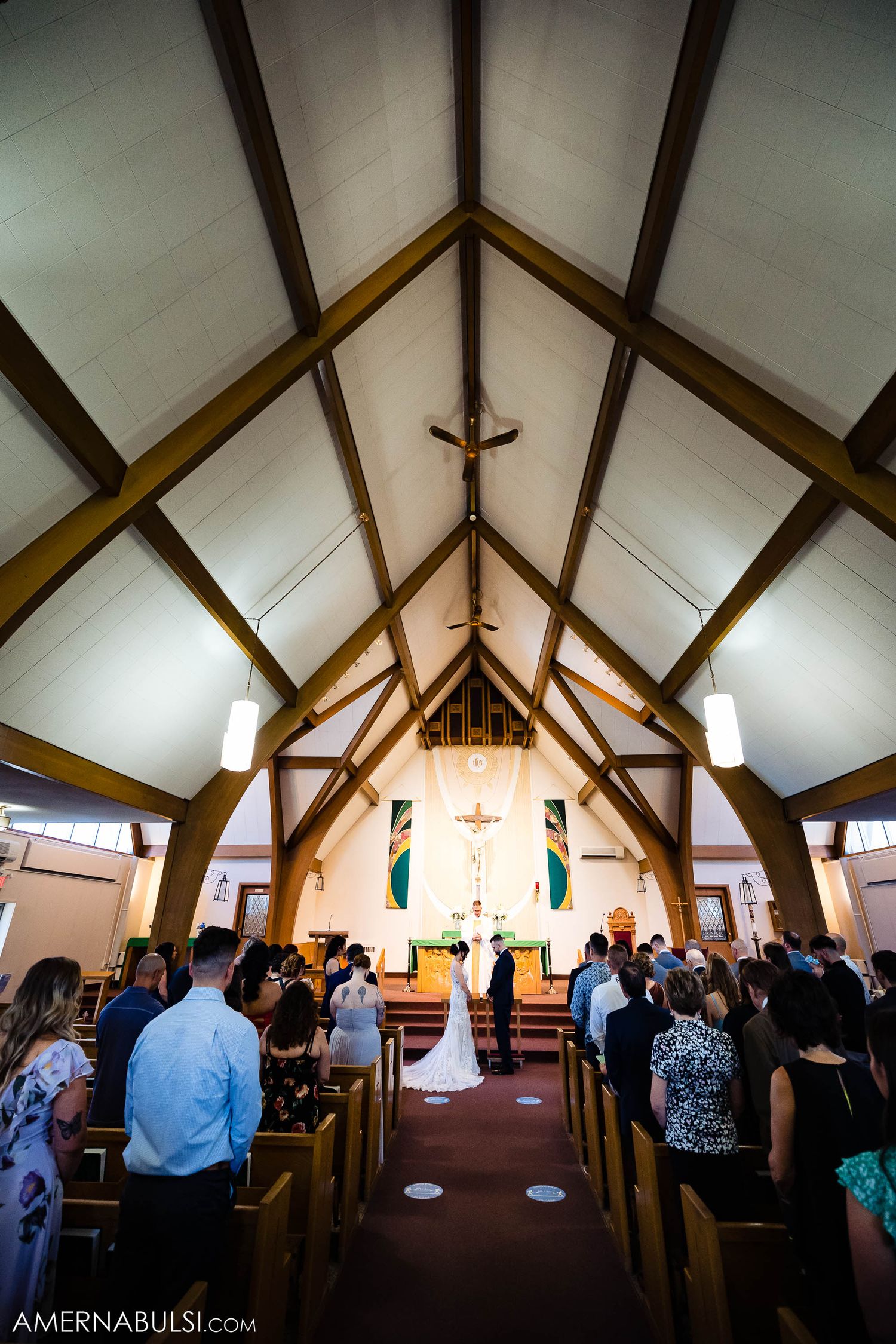 Church interior with wooden beams and vaulted ceiling during a wedding ceremony with guests in the pews.