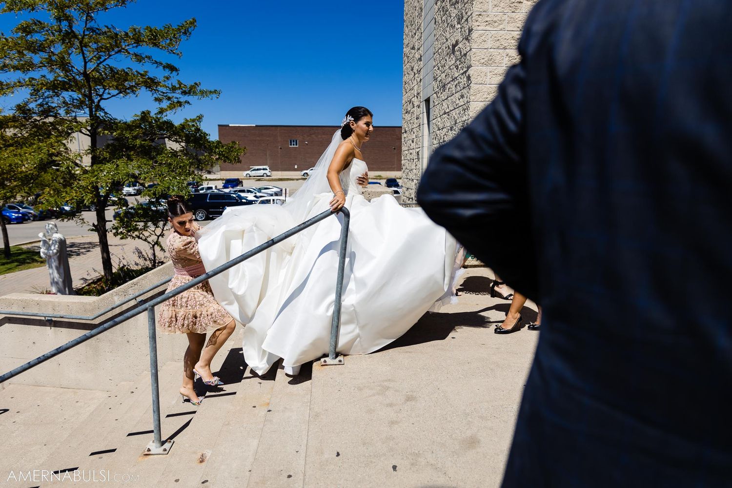 Bride in white dress carefully descending outdoor stone steps with assistance.