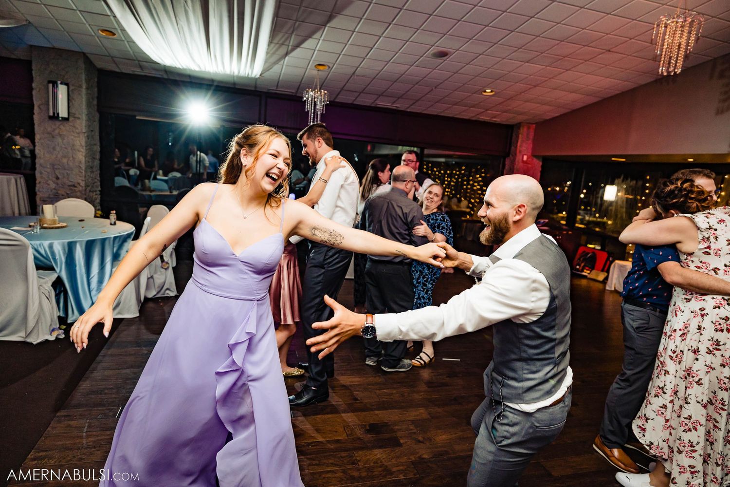 Wedding guests dancing energetically on a dark wooden dance floor at an indoor reception venue.
