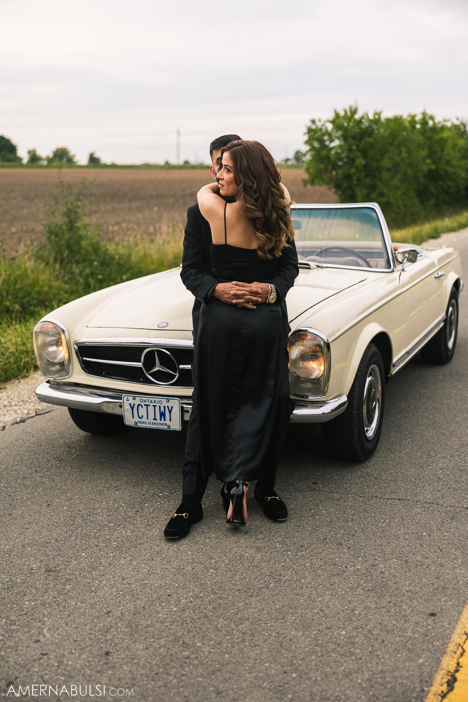 Elegant couple embraces in front of white vintage Mercedes-Benz convertible.