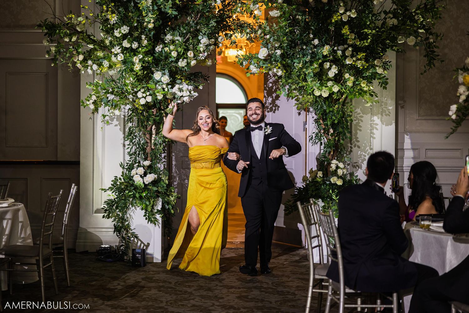 Wedding guests in formal attire entering reception venue under decorative floral arch.