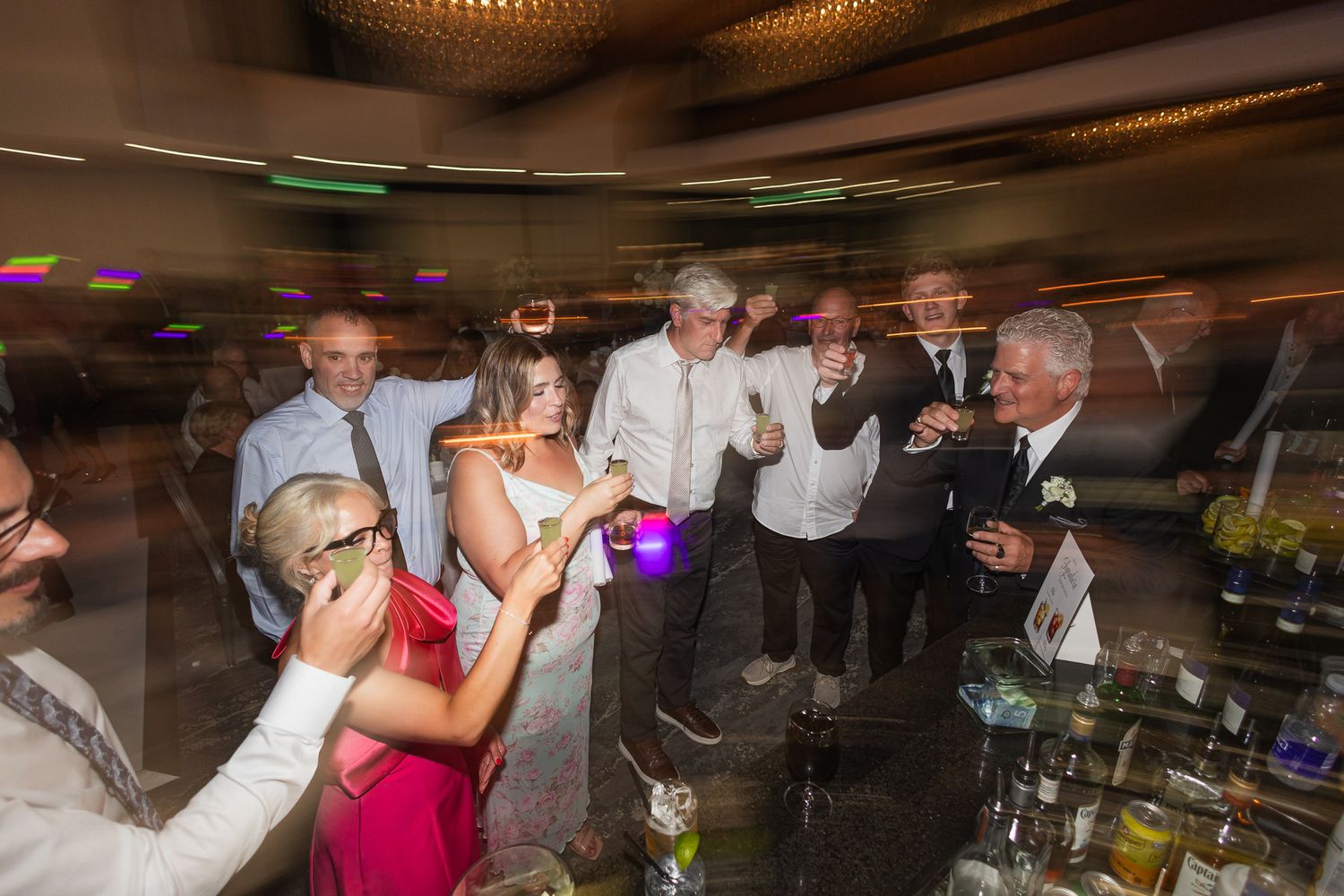 Guests raising glasses in a toast at a lively wedding reception party near a bar.