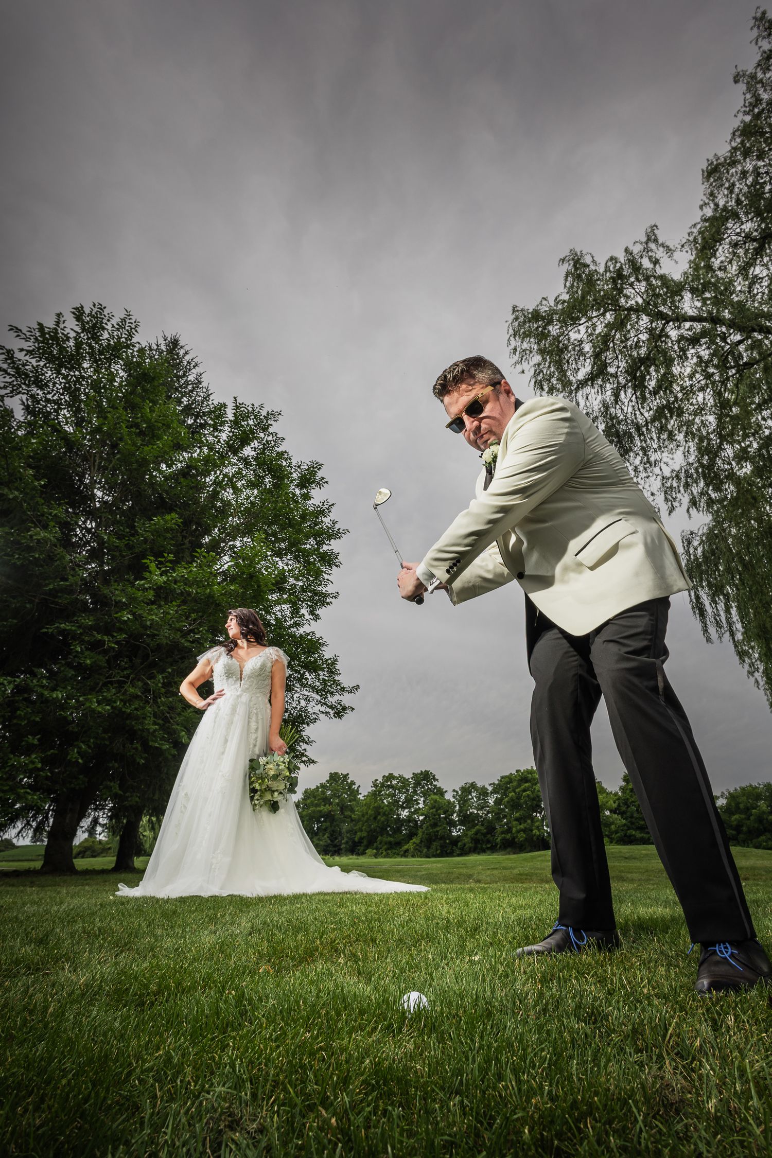 Groom in sunglasses swings golf club on green lawn while bride in white gown holds bouquet on their wedding day.