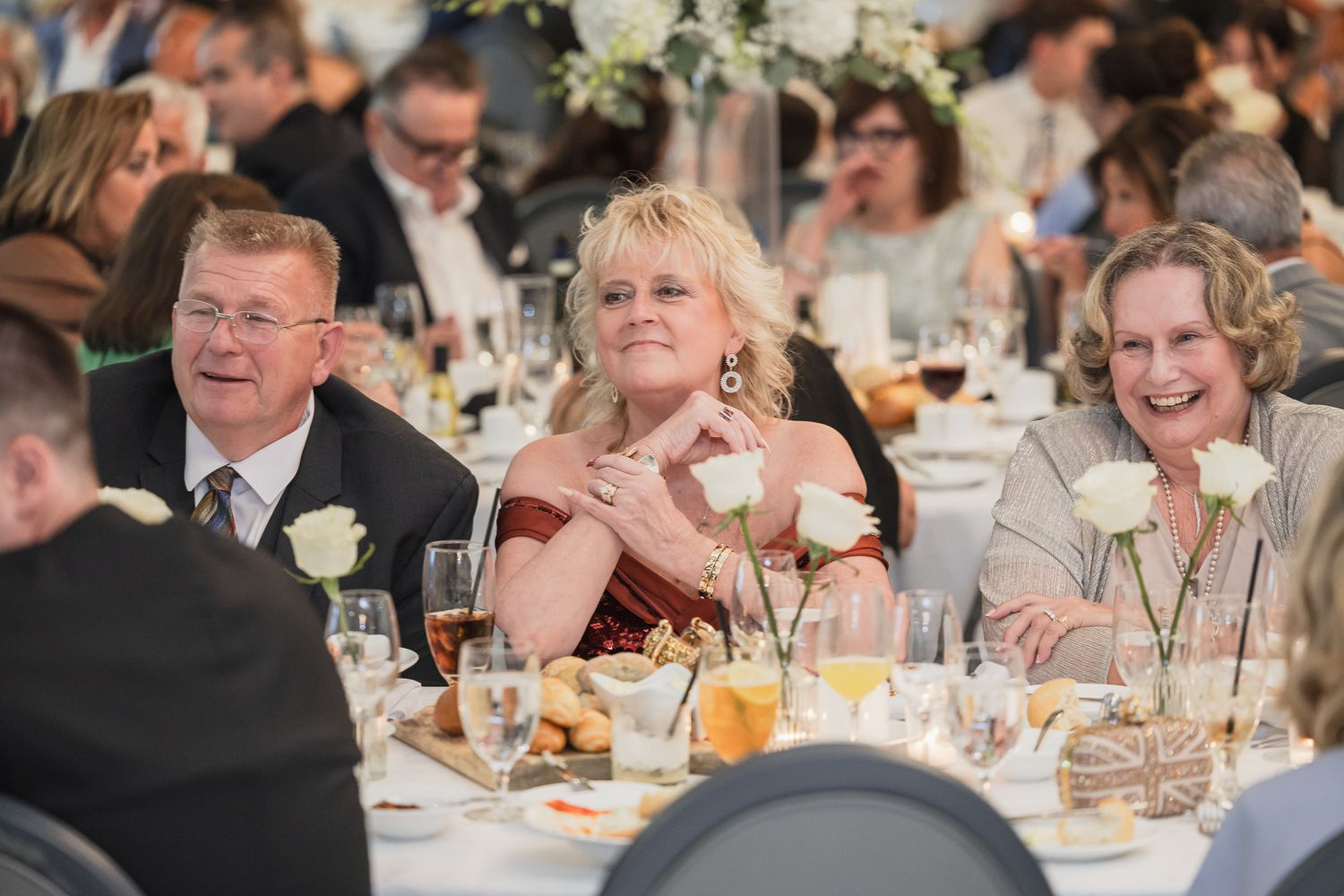Three wedding guests smile and laugh at a decorated reception table with white roses and glassware.