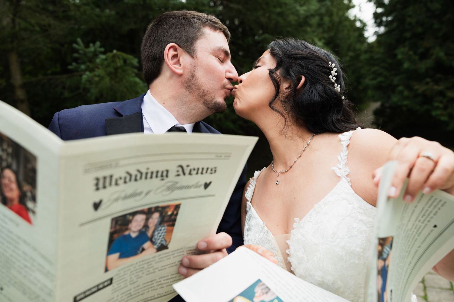 Bride and groom sharing a kiss while holding a personalized Wedding News newspaper at their wedding.