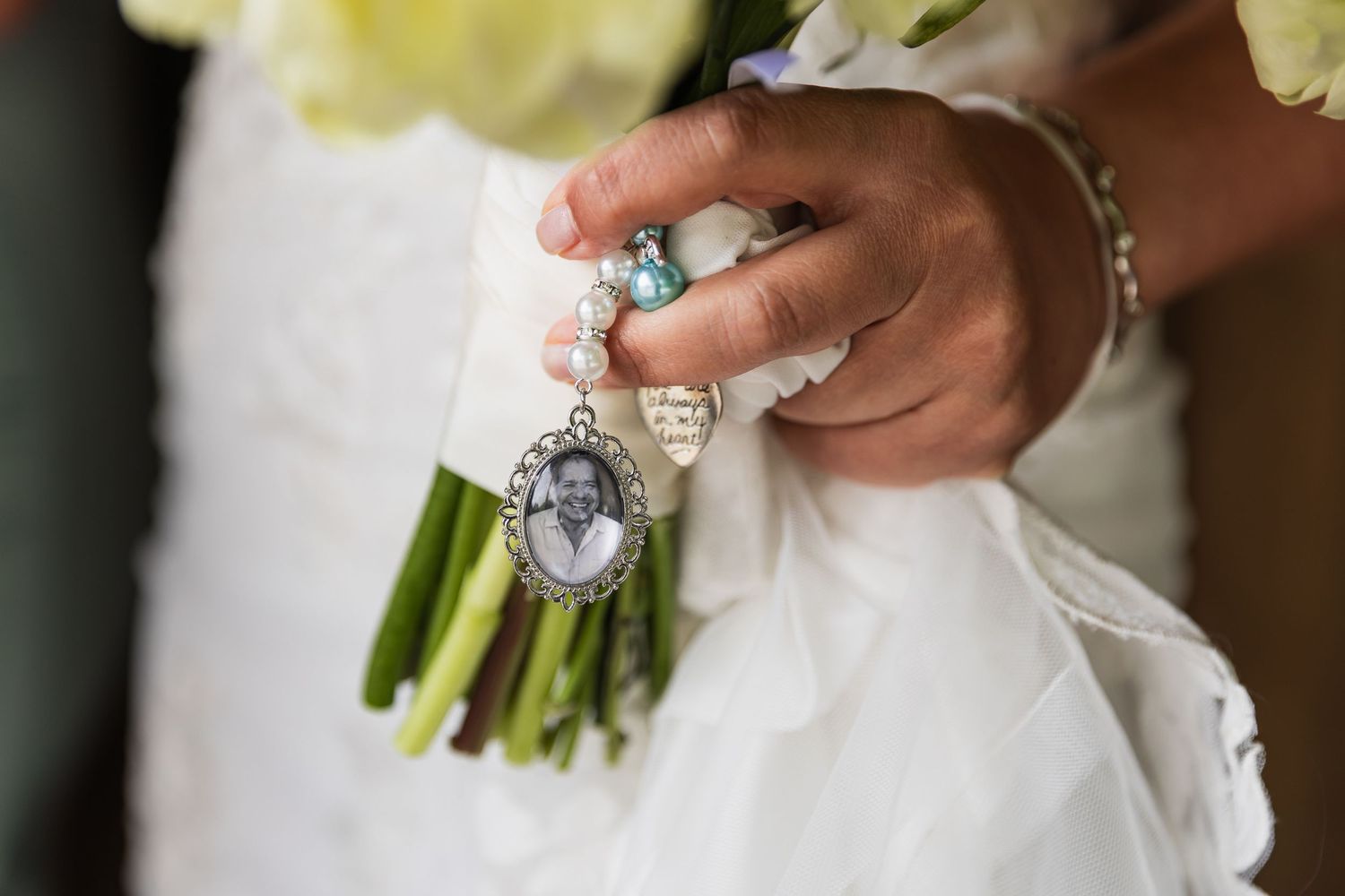 Bride holding bouquet with memorial photo charm and pearl bracelet as a tribute to loved one on wedding day.