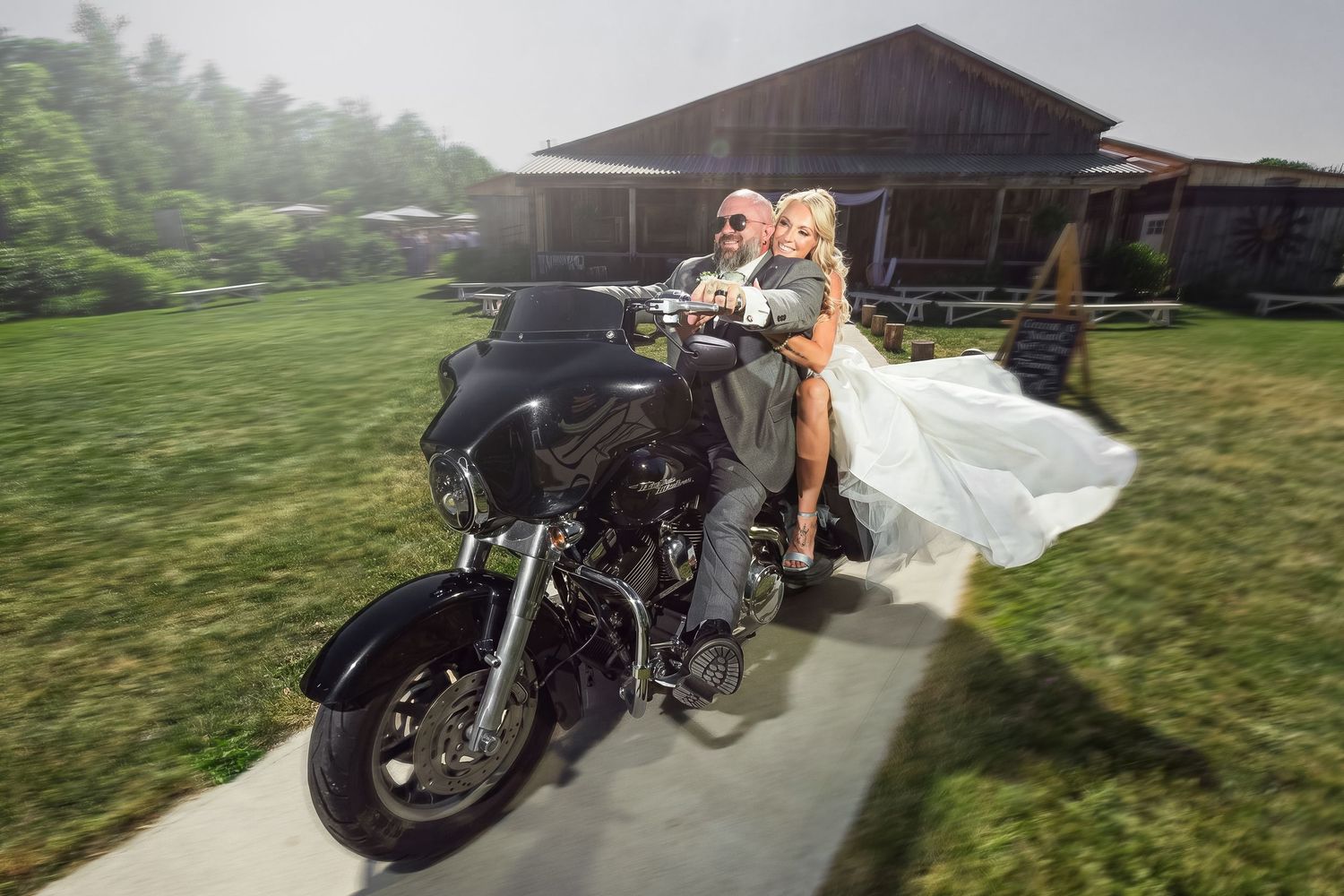 Bride and groom ride Harley-Davidson motorcycle outside rustic barn venue on their wedding day.