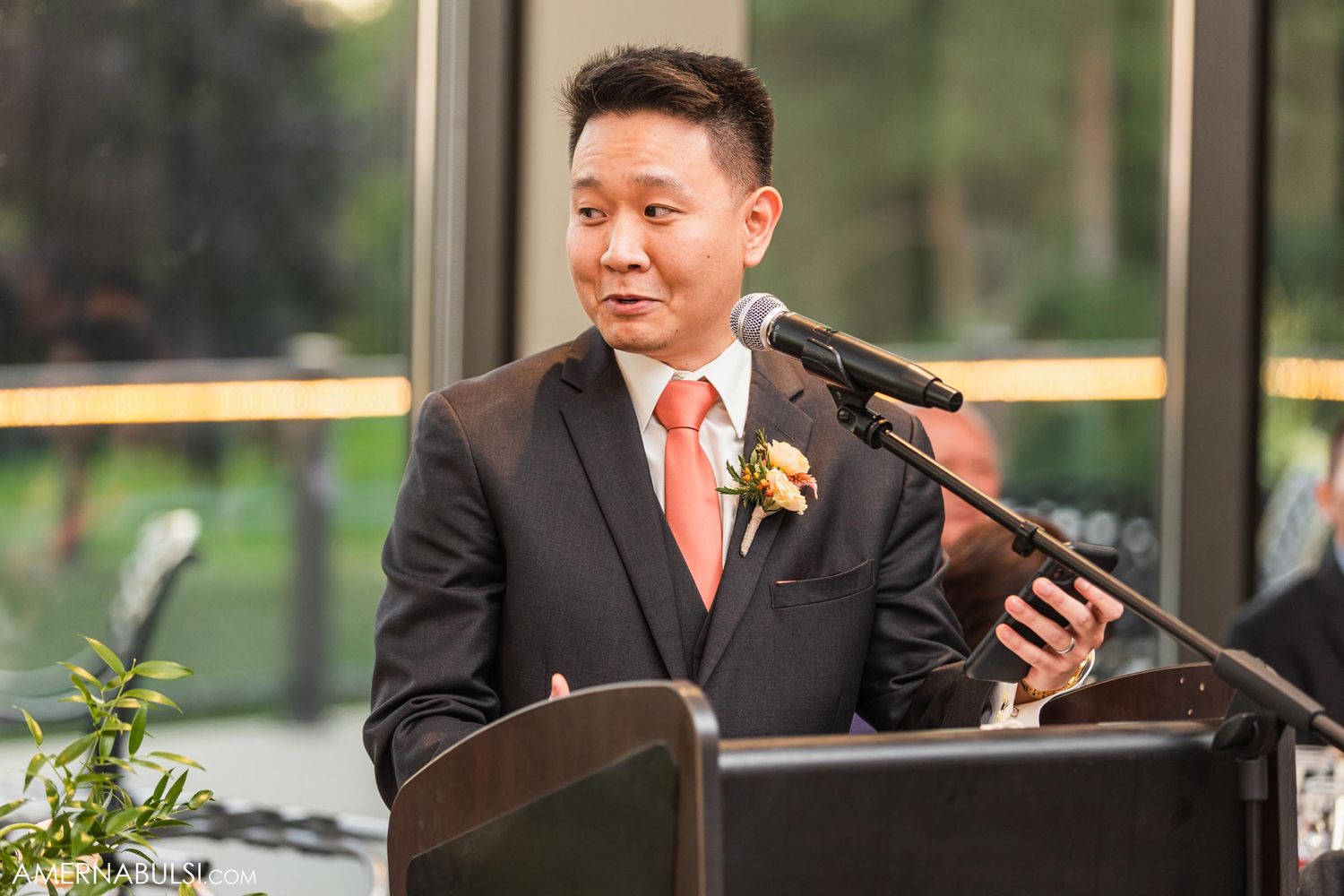 Groom delivering speech at wedding reception podium wearing dark suit with coral tie.