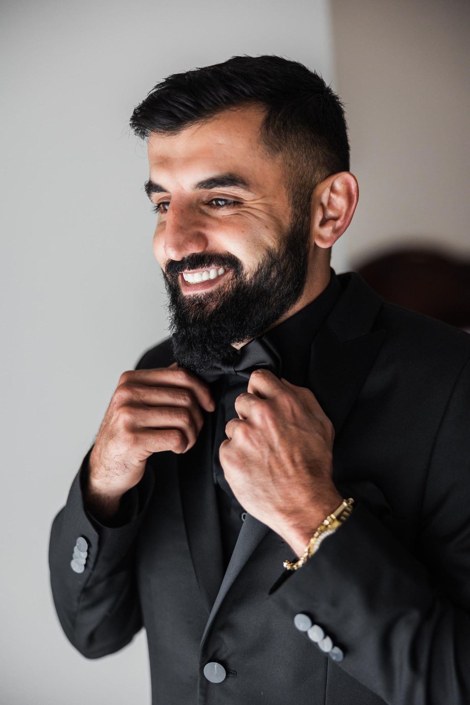 Stylish groom adjusts his black bow tie while smiling in formal wedding attire.