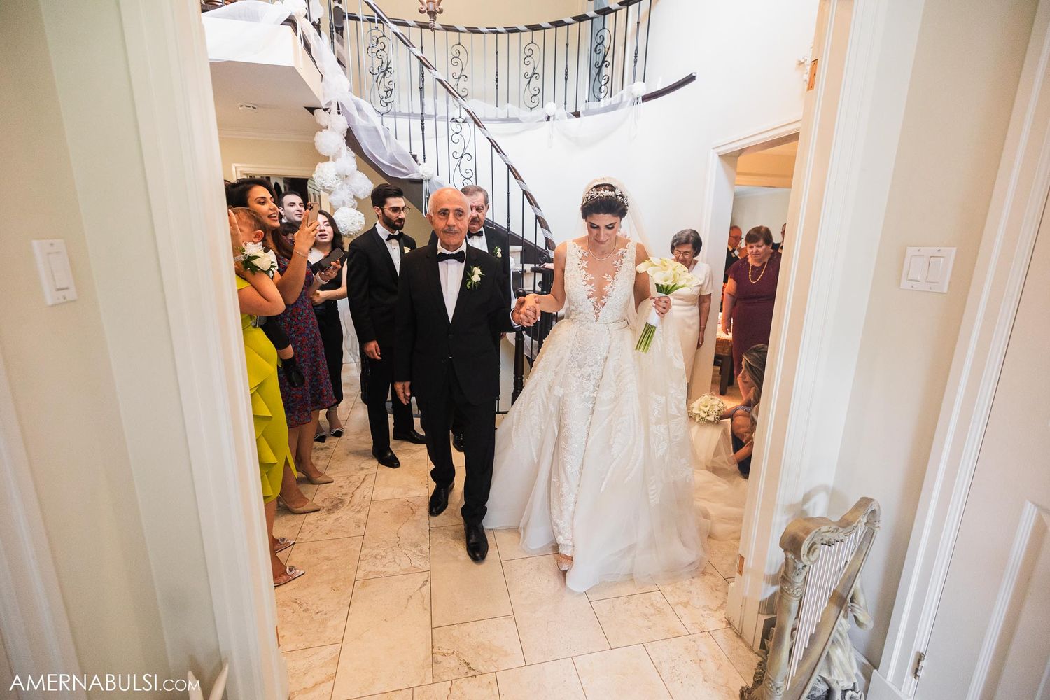 Wedding party walks through marble foyer with elegant staircase in background.