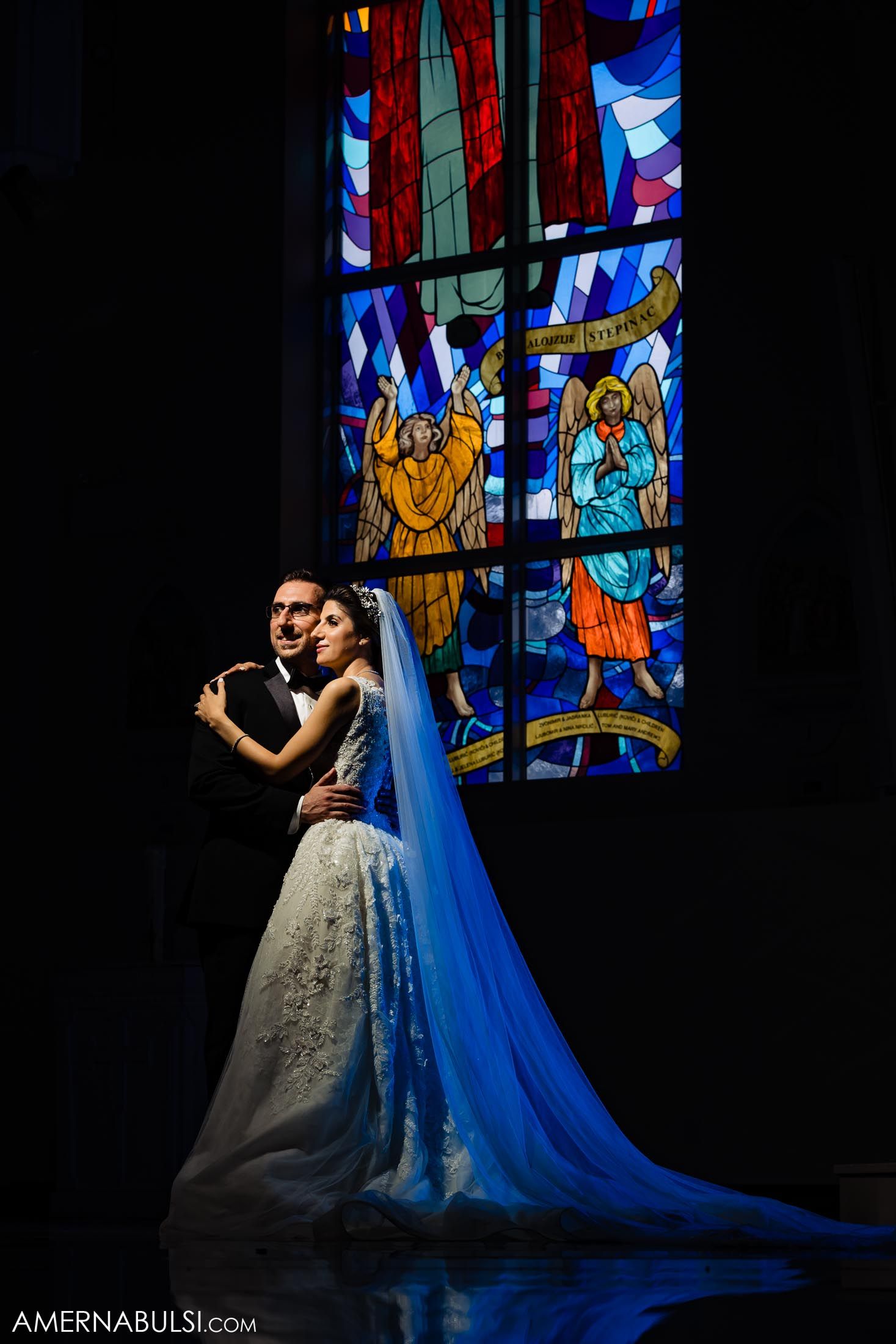 Wedding couple embraces in dramatic lighting against colorful stained glass church window.
