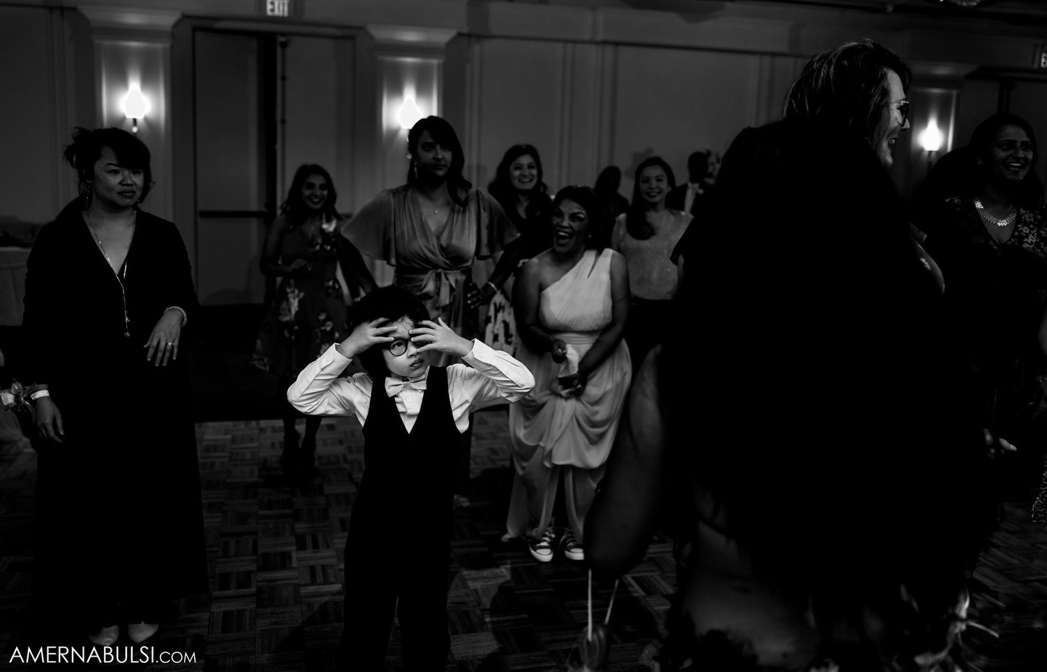 A dramatic black and white photograph of wedding guests dancing in a dimly lit reception hall.