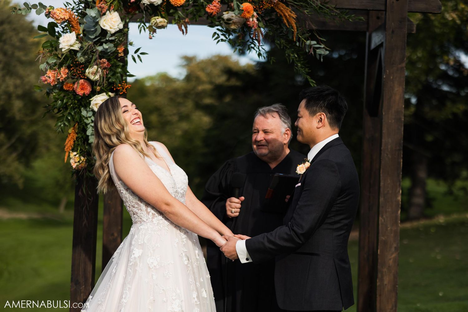 Wedding ceremony under a wooden arch decorated with orange and white flowers in a garden setting.