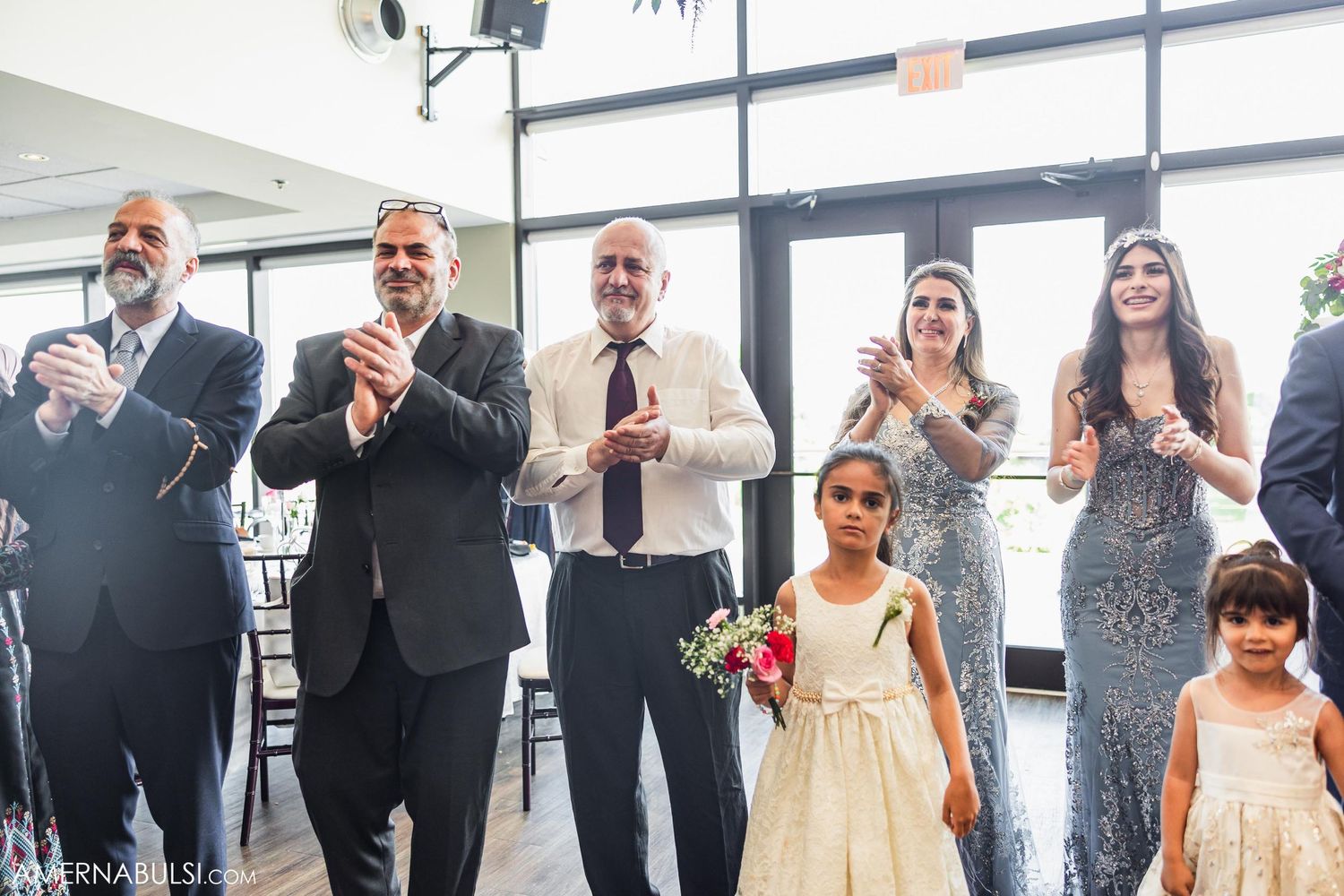 Wedding guests and children in formal attire applaud during reception festivities.