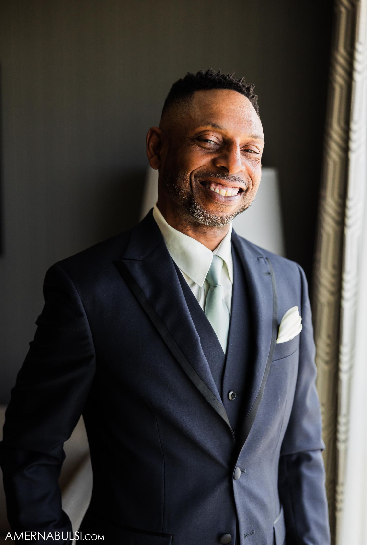 A groom in a navy suit and mint green tie poses by a window for a formal portrait.