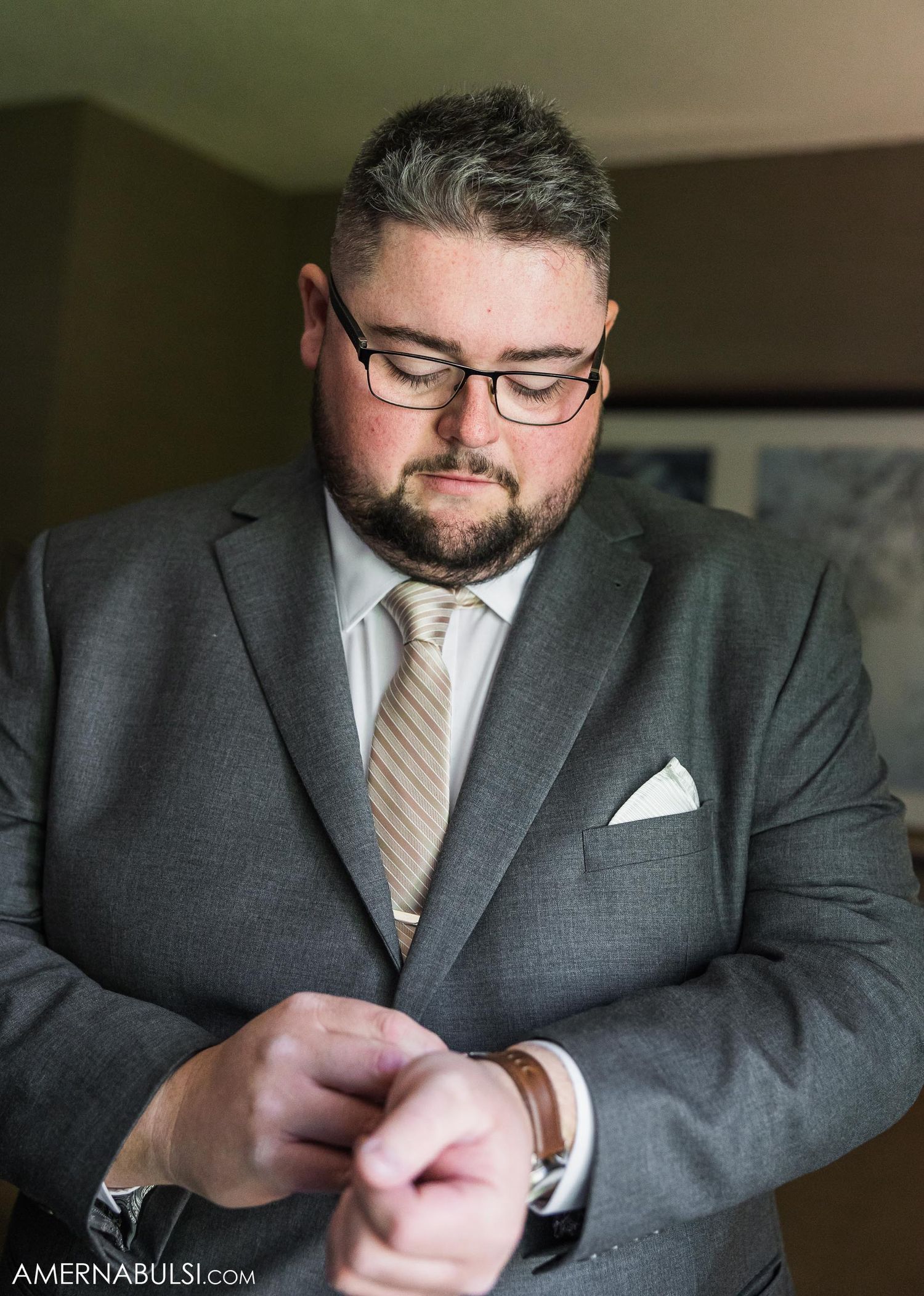 Groom in grey suit and glasses adjusting cuffs during wedding preparation.