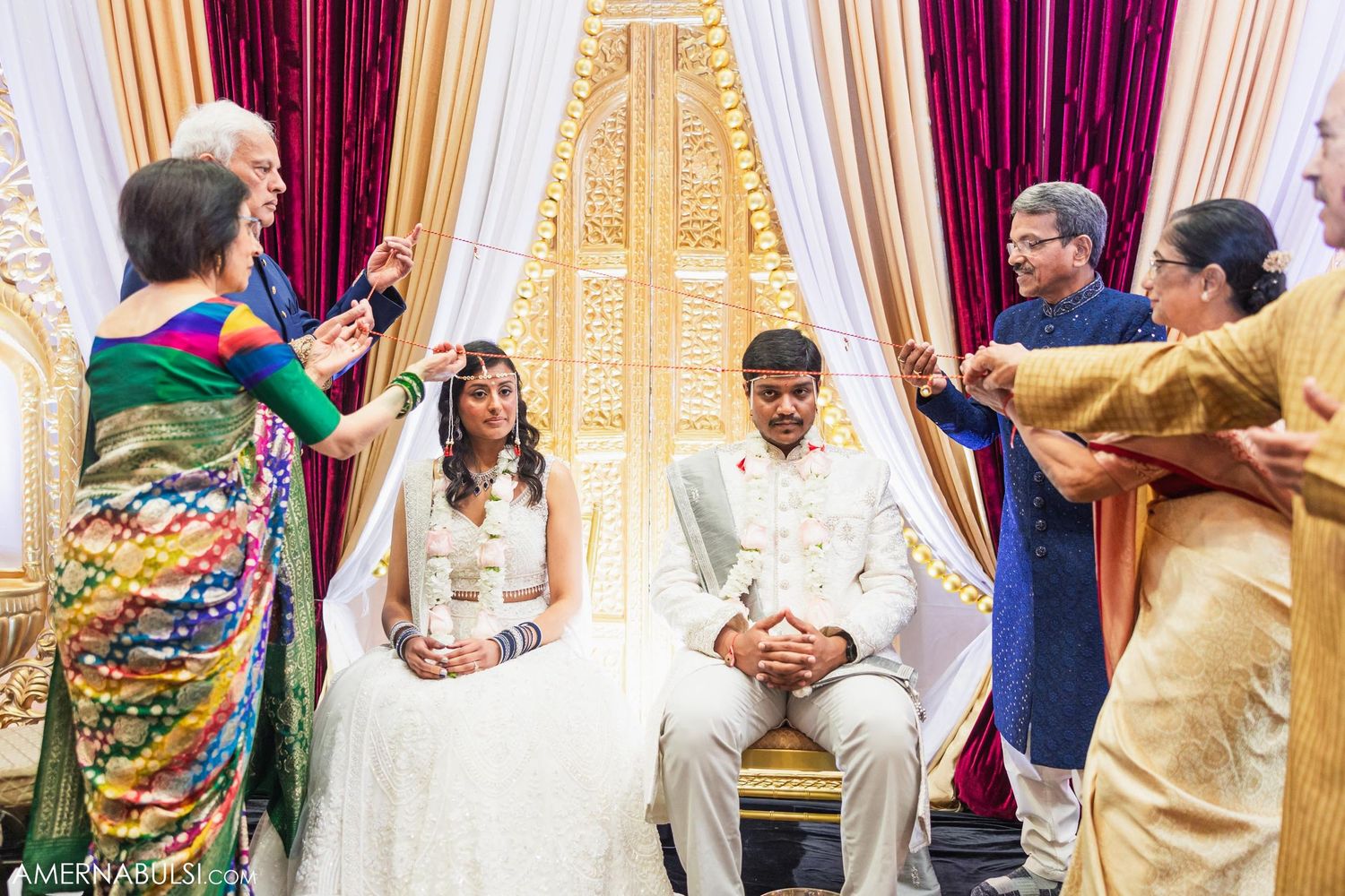 Wedding ceremony attendees performing traditional rituals with colorful attire and white drapery.