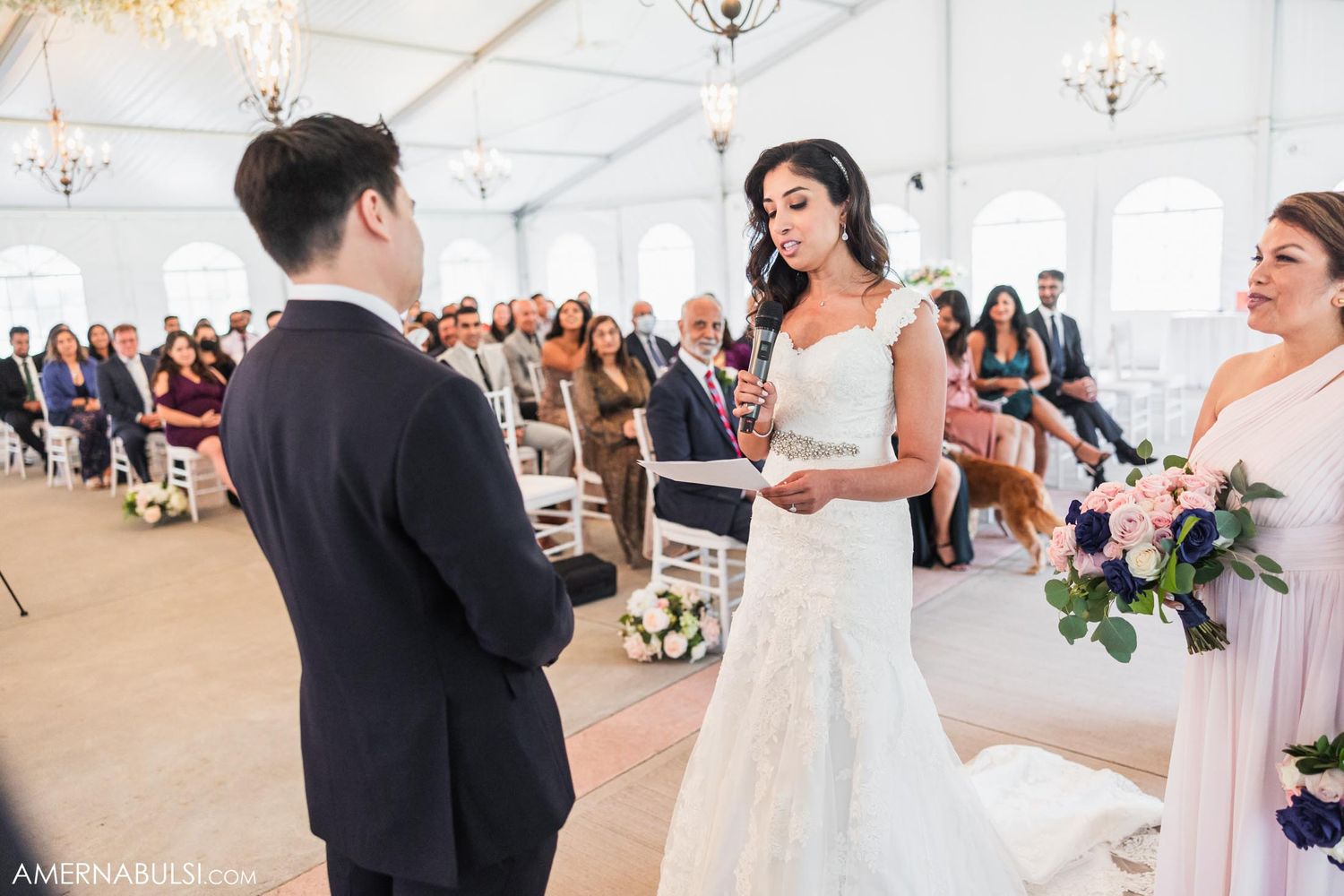 Wedding ceremony in progress in an elegant white tent with crystal chandeliers.