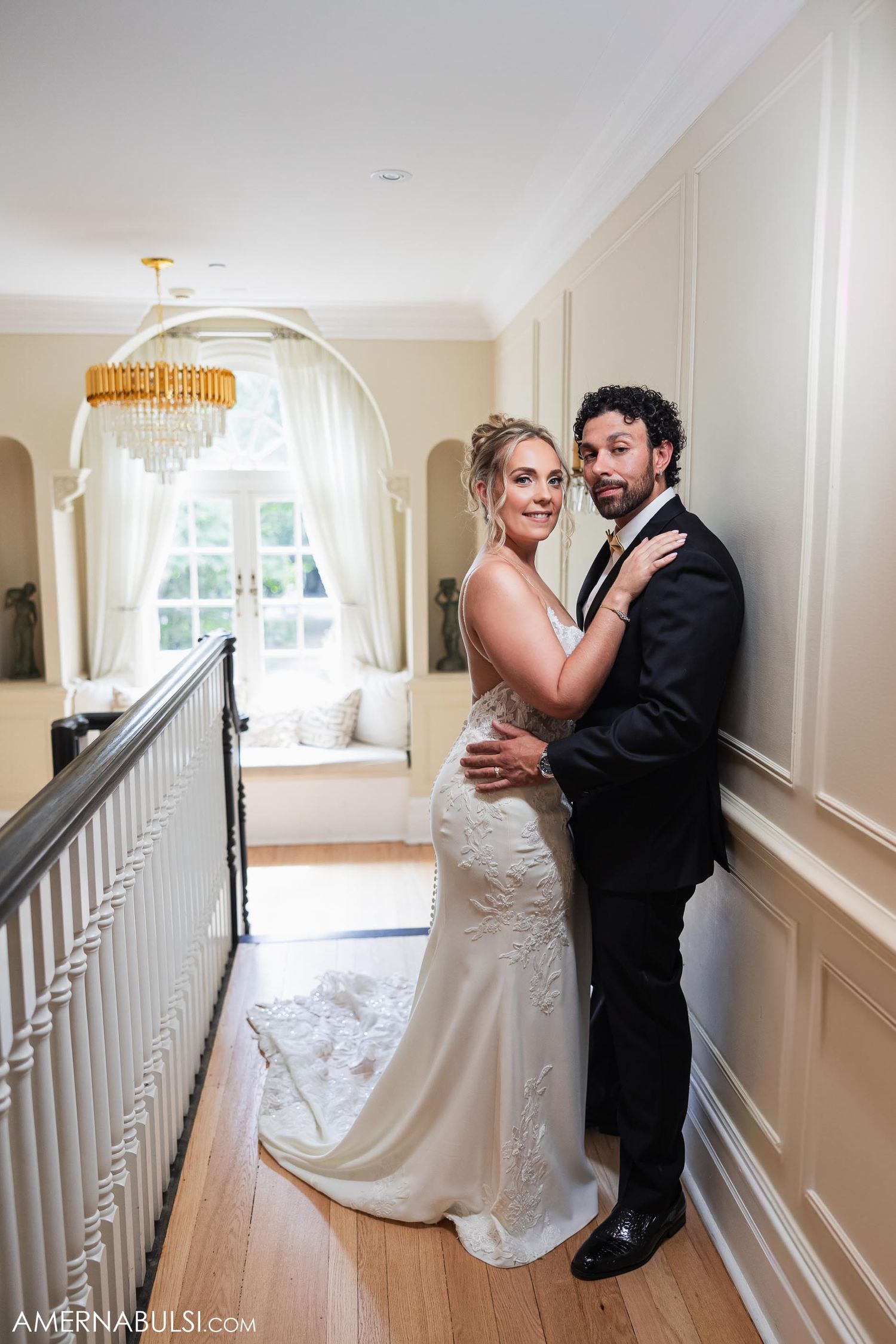 Wedding couple embraces in an elegant hallway with crystal chandelier and white interior details.