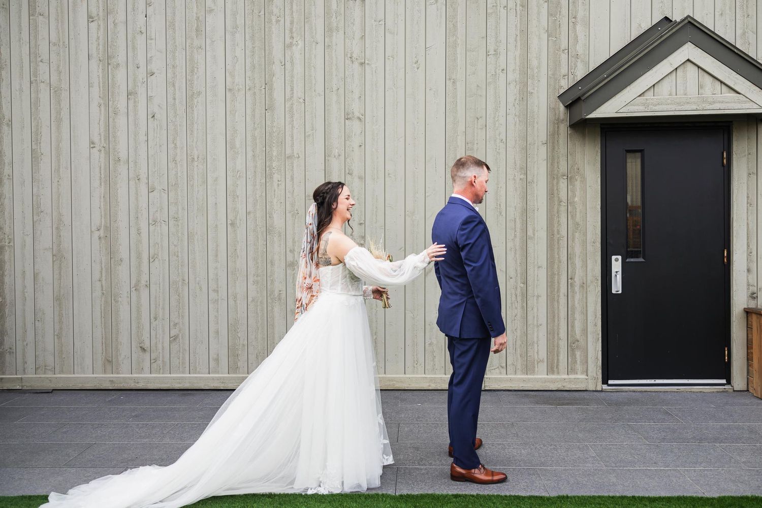 First look wedding moment with bride and groom against grey building exterior.