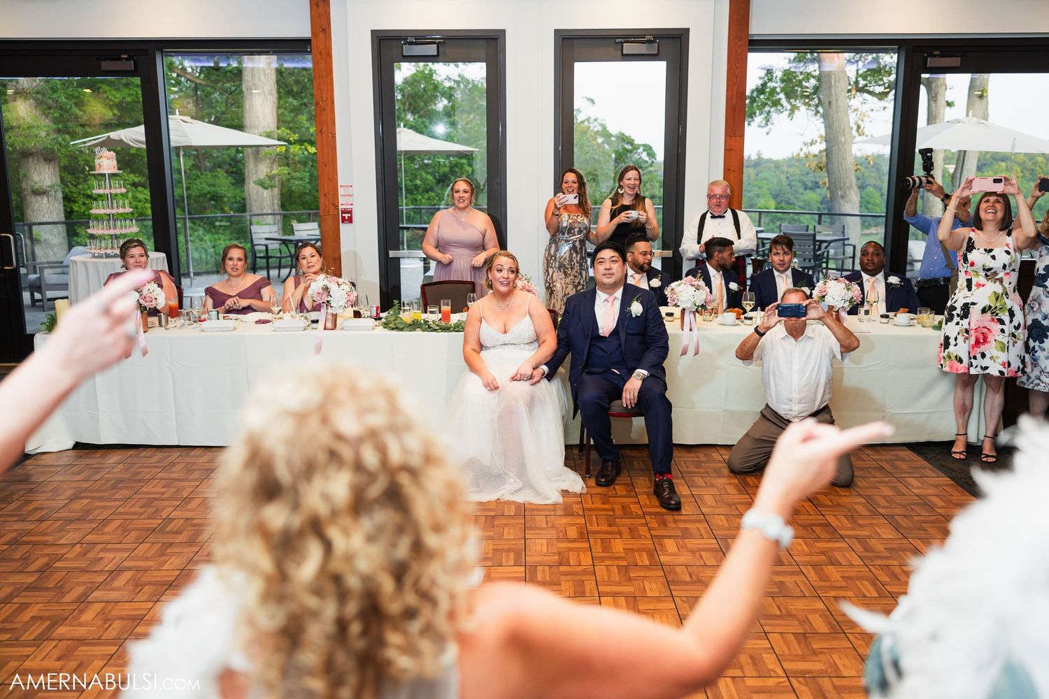 Wedding guests watch and take photos during reception celebration in venue with large windows.