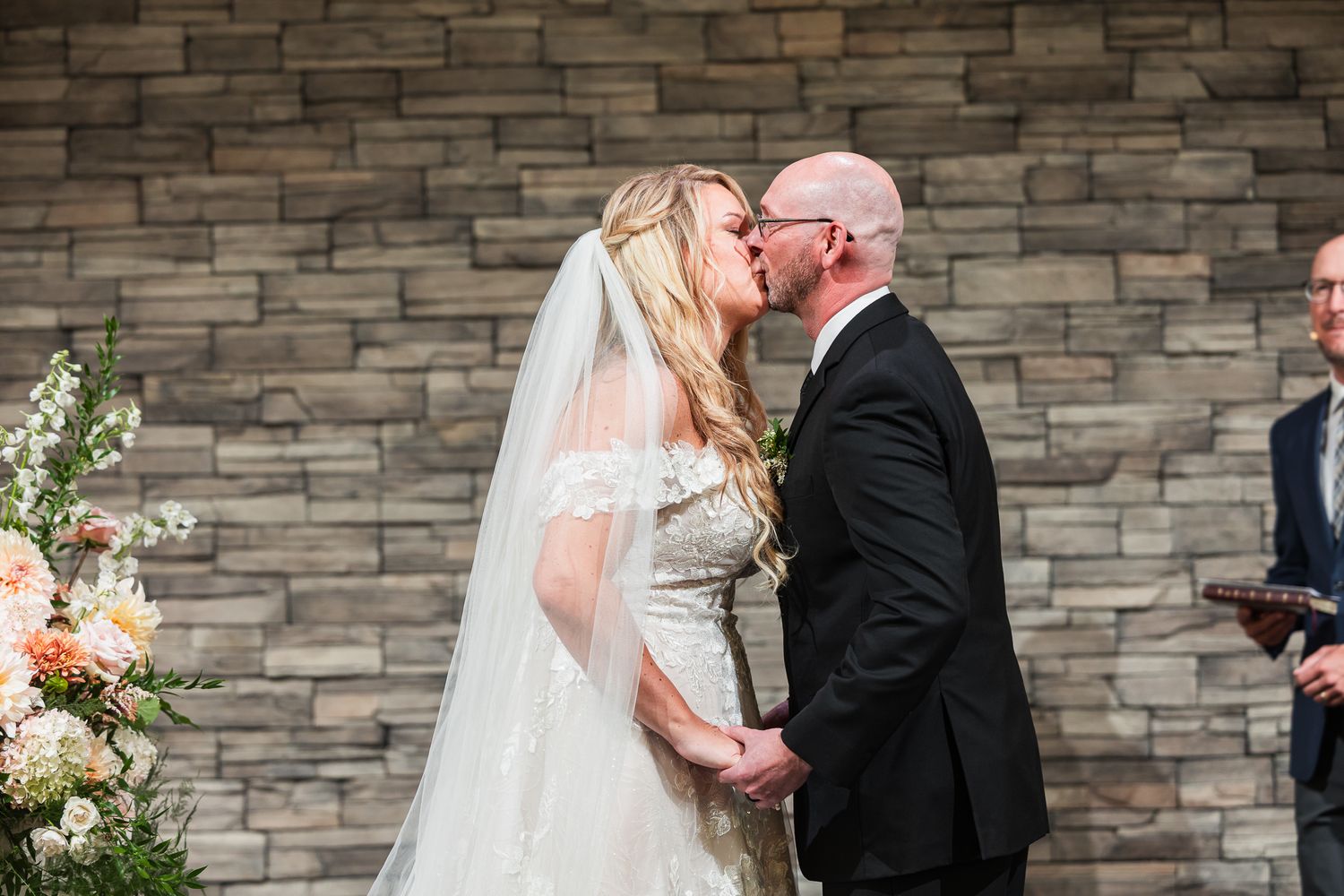 A tender wedding kiss moment against a stone wall backdrop with elegant floral arrangements.