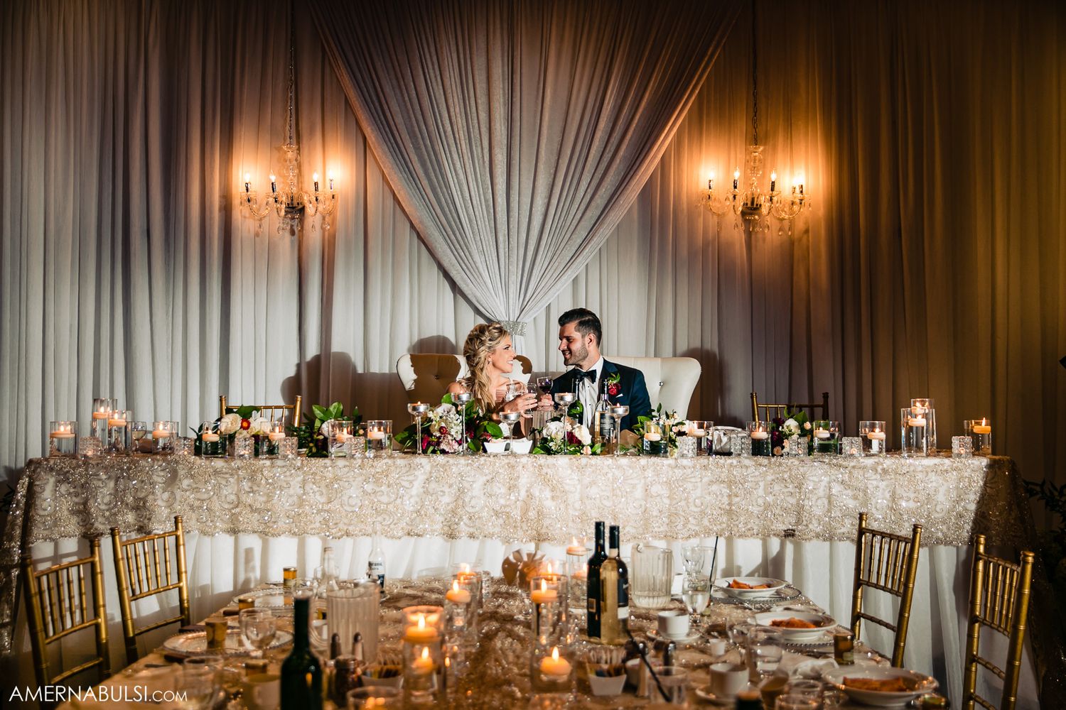 A panoramic view of an elegant wedding reception hall with white draping, chandeliers and decorated dining tables.