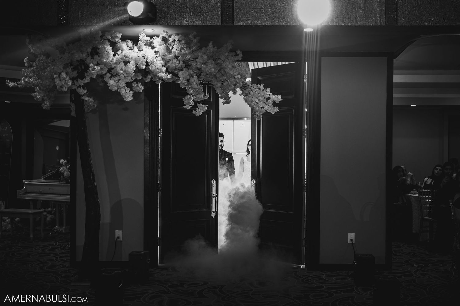 Atmospheric black and white photo of a wedding venue entrance with floral decorations and dramatic lighting.