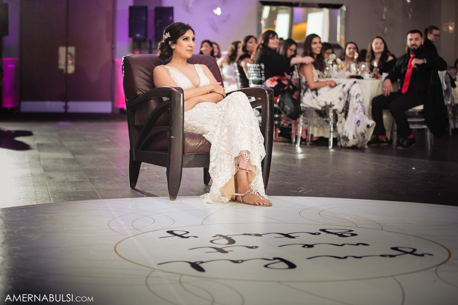 A wedding reception scene showing someone in white seated in a chair on a dance floor with guests in the background.