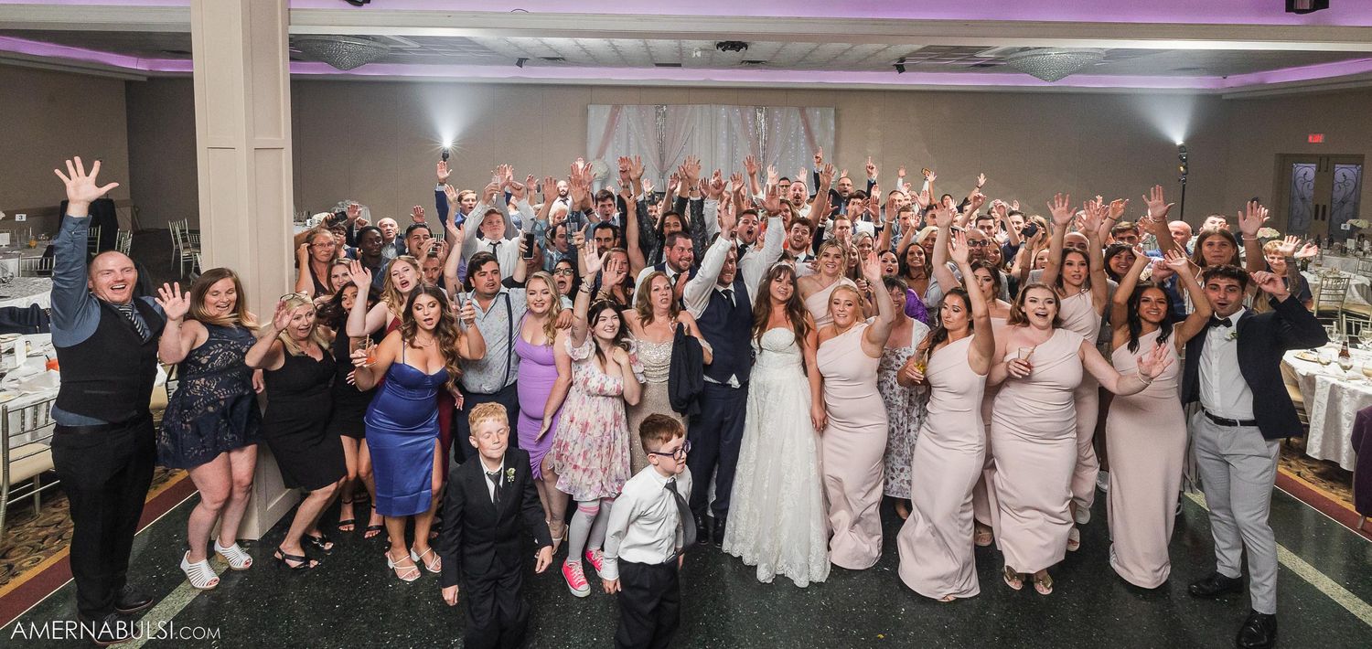 Large wedding group photo with guests raising their hands in celebration.