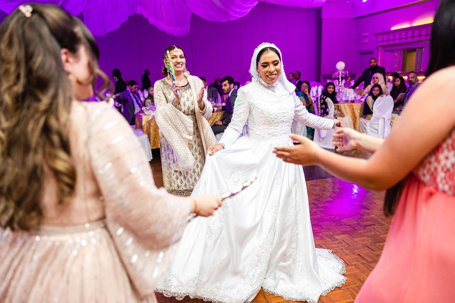 Bride in white dress dances with guests during reception under vibrant purple lighting.