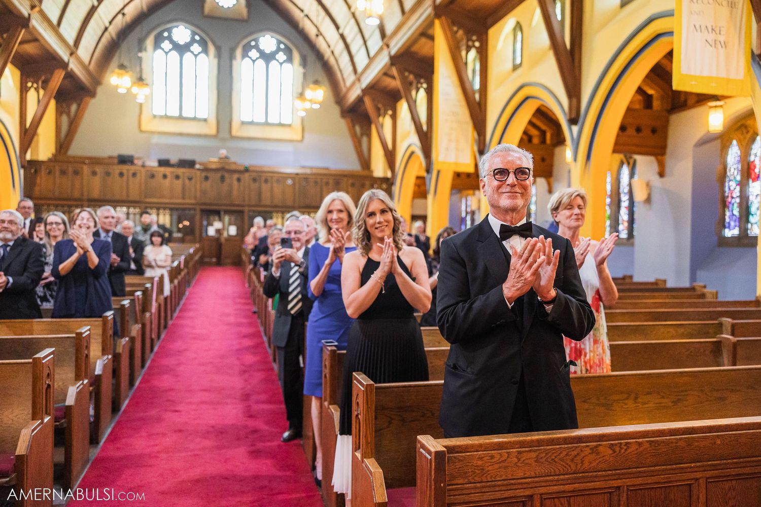 Wedding guests applaud while standing in wooden pews inside a grand church with vaulted ceilings.