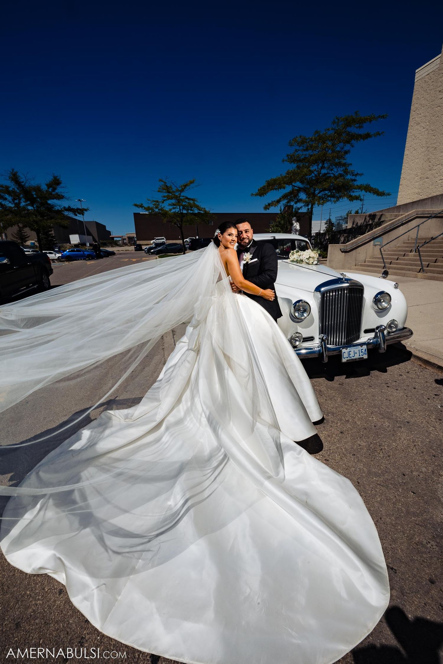 Classic white wedding dress with long flowing train posed against vintage Rolls Royce under blue sky.