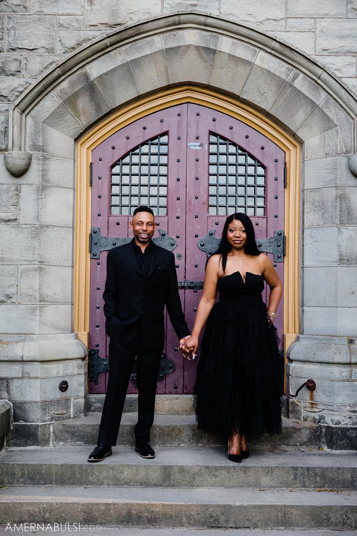 Couple in formal black attire poses in front of ornate pink gothic doorway.
