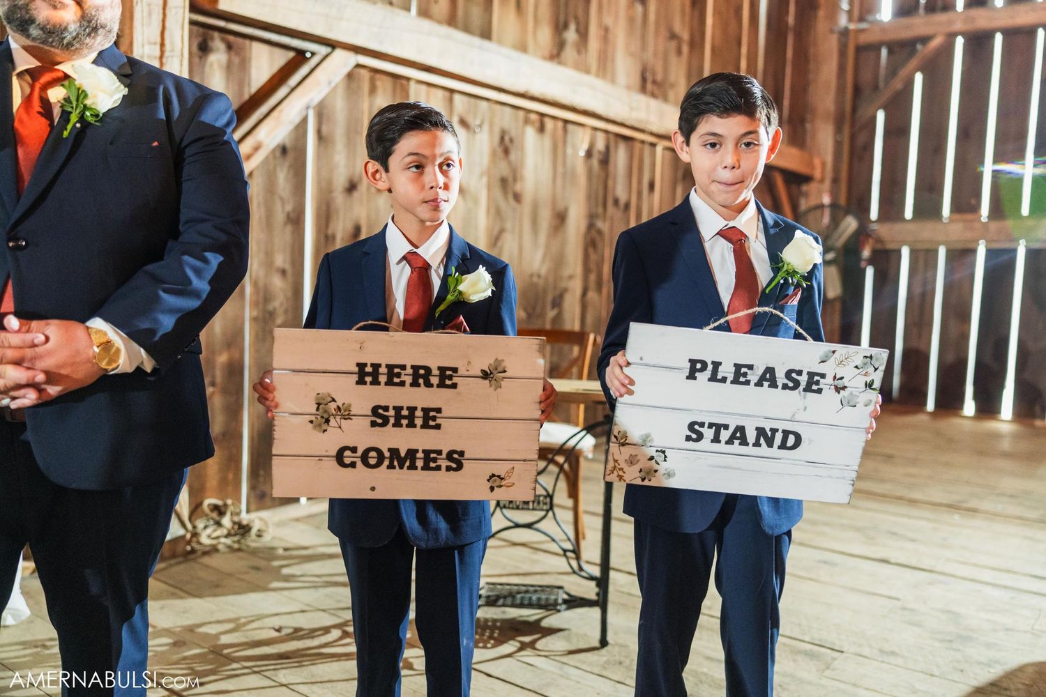 Two young attendants hold wooden signs reading 'Here she comes' and 'Please stand' in rustic barn setting.