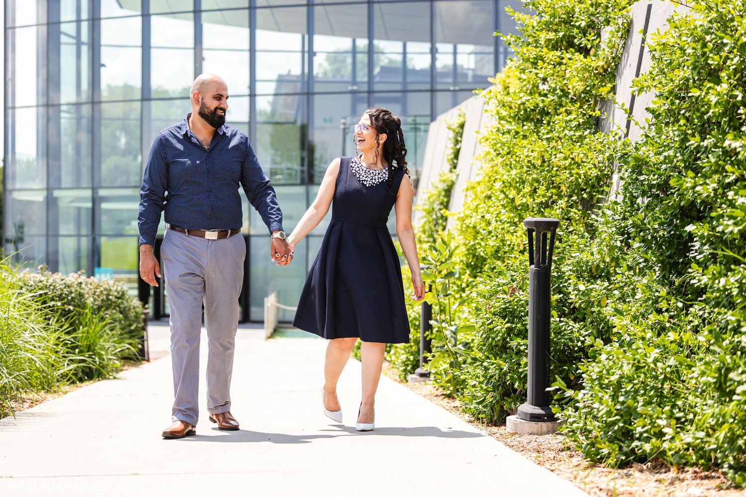 A couple strolls hand in hand along a sunny garden pathway beside a modern building.