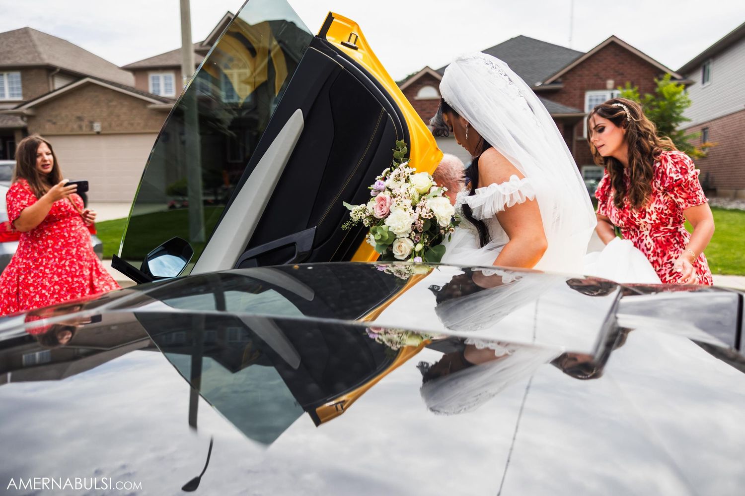 Wedding guests in red dresses watch as bride looks into yellow sports car.