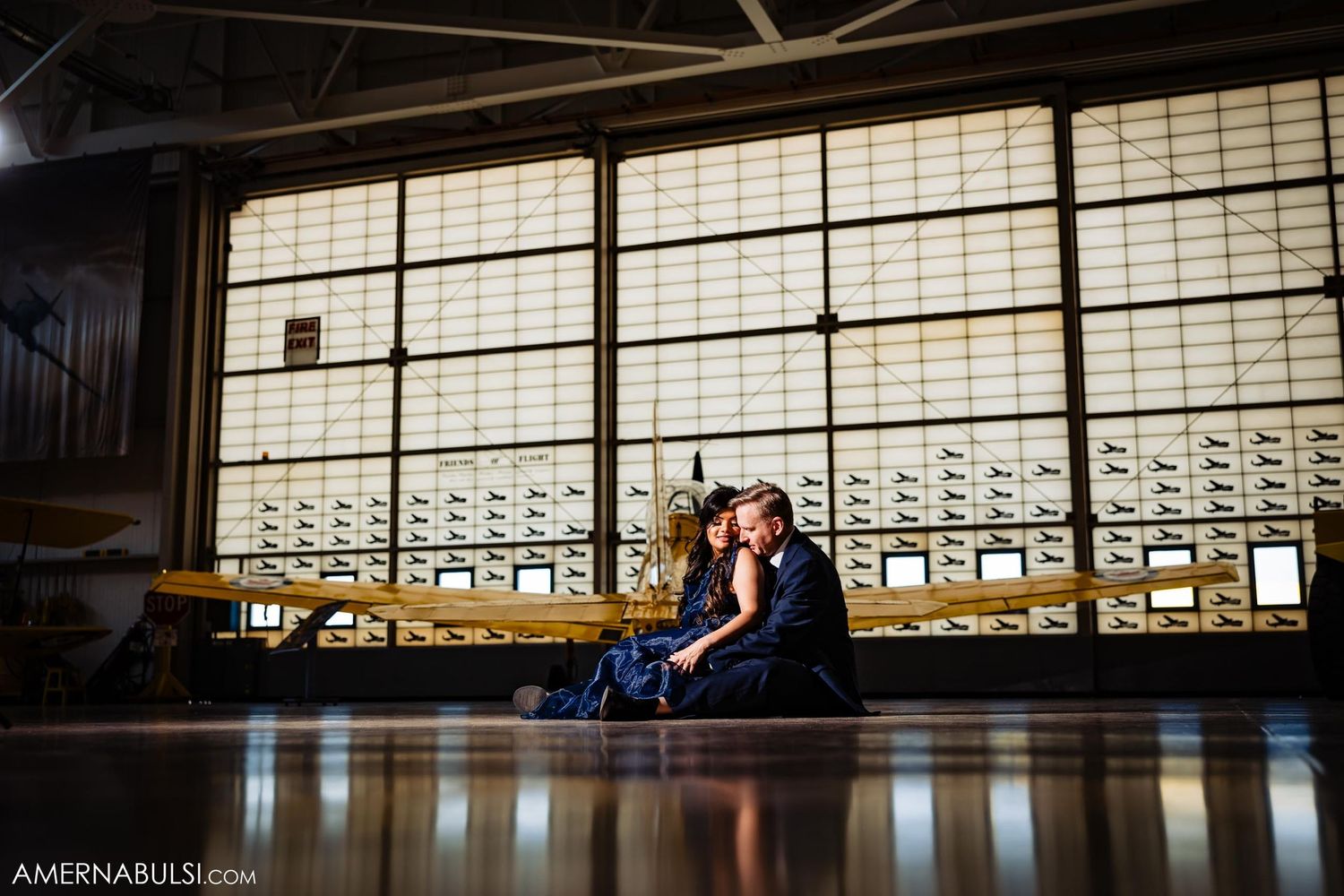 A couple sits together in a dimly lit hangar with window light streaming in.