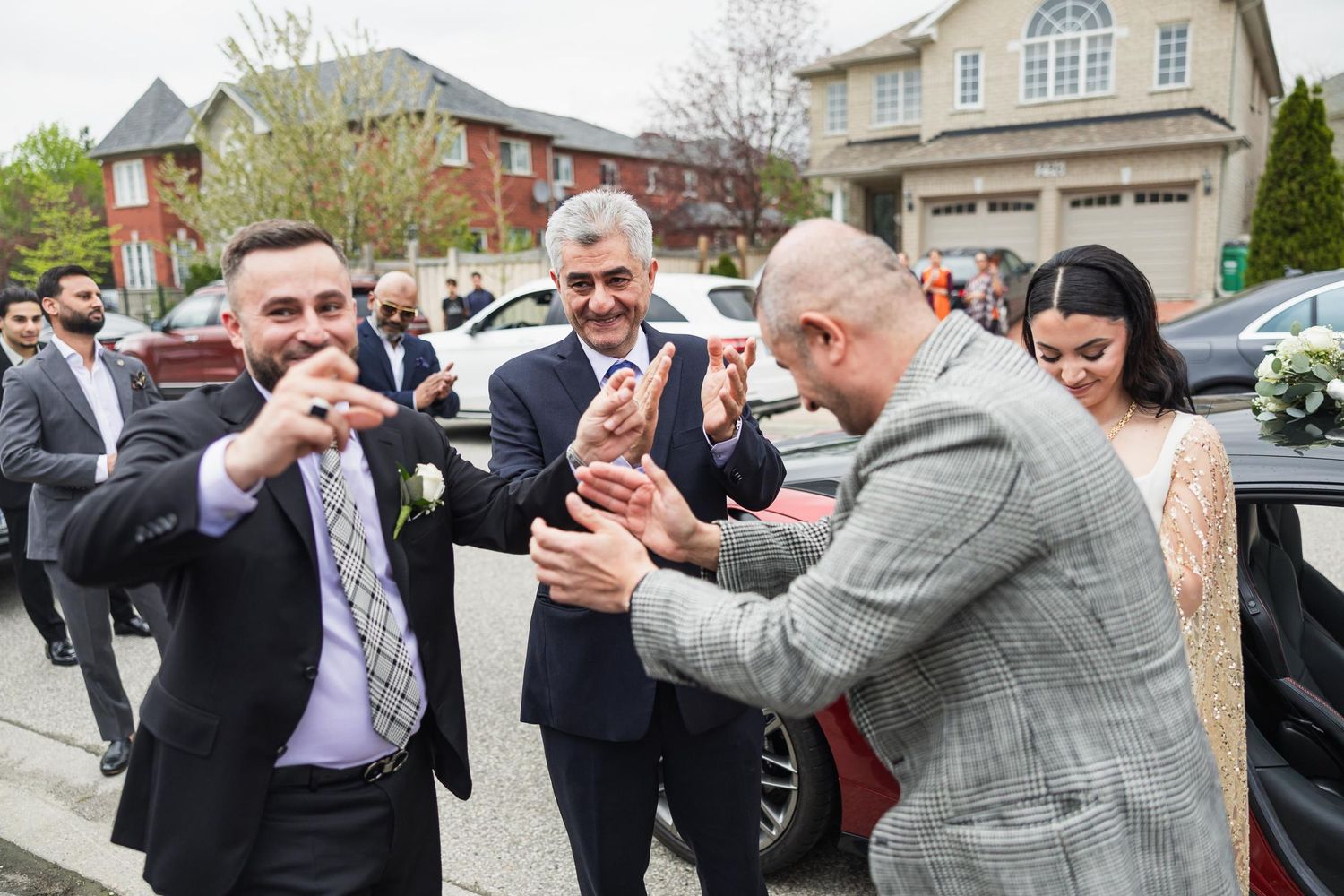 Wedding guests celebrate by dancing and clapping together outside a suburban home.