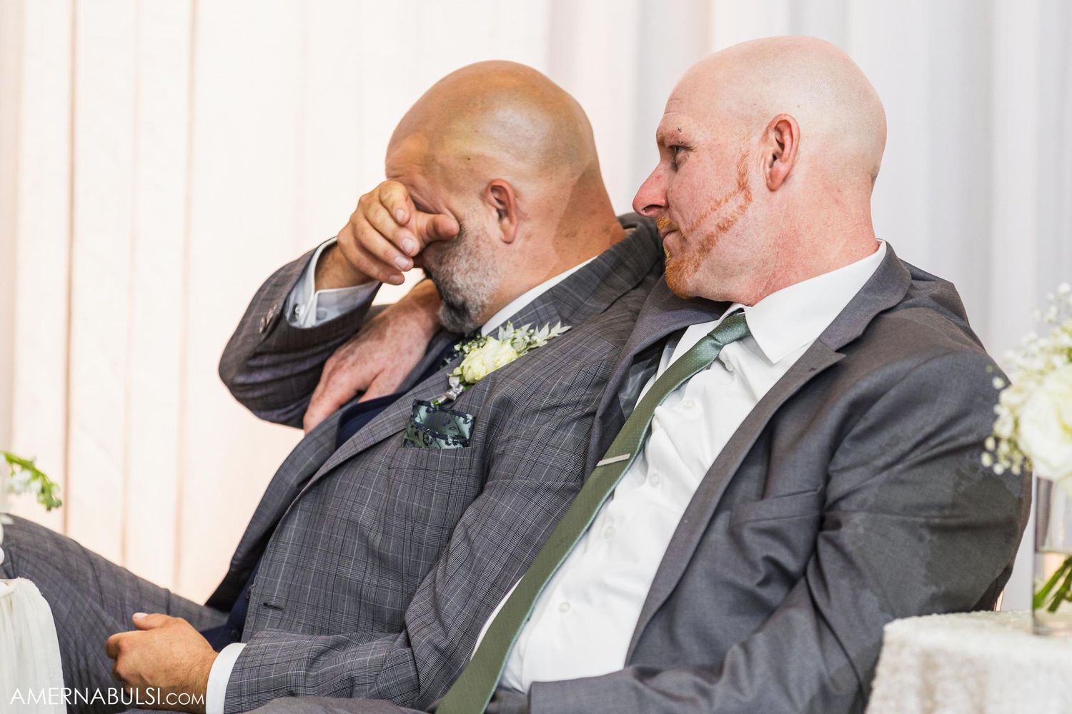Two men in grey suits laughing together at formal event with boutonnières.