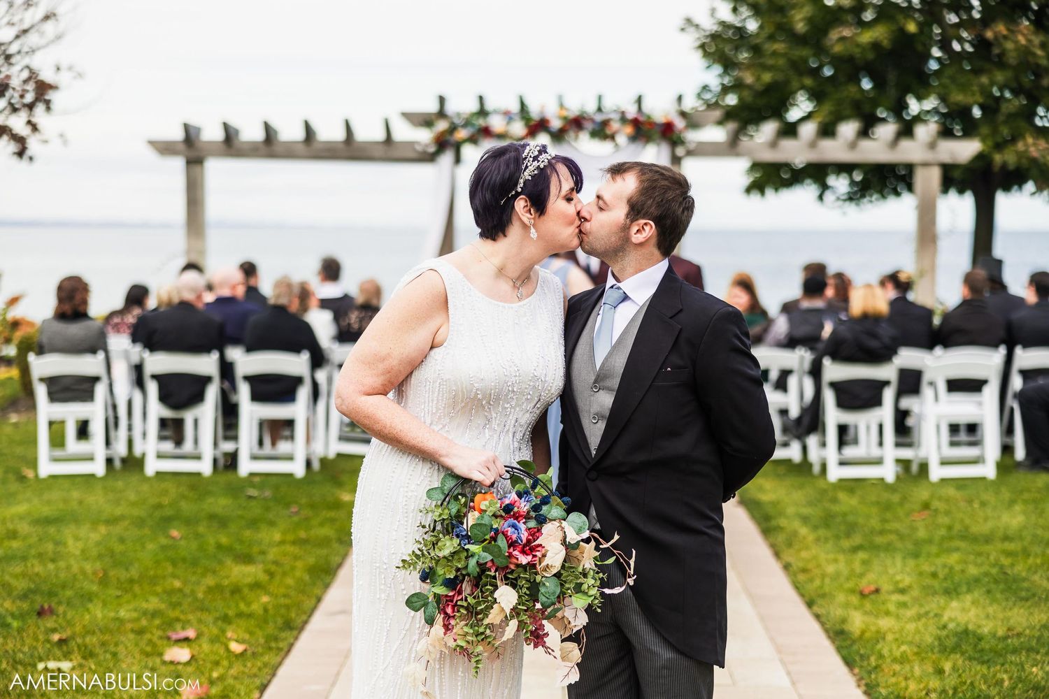 Waterfront wedding ceremony with white chairs and wooden pergola overlooking the ocean.