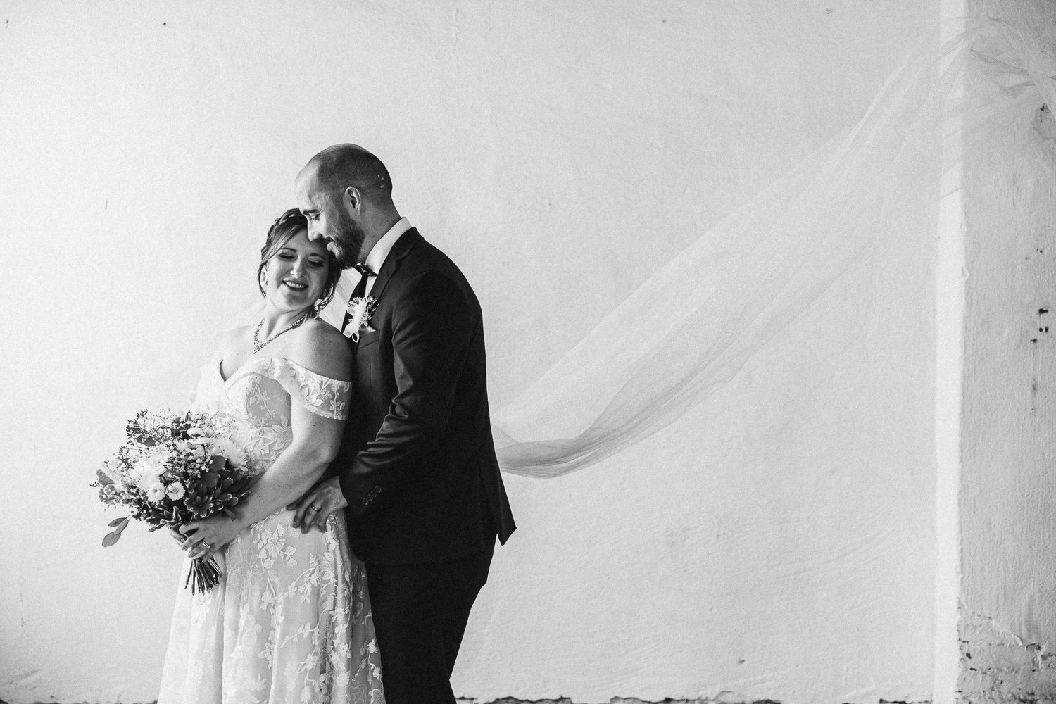 A romantic black and white wedding portrait with couple holding a floral bouquet against a white backdrop.