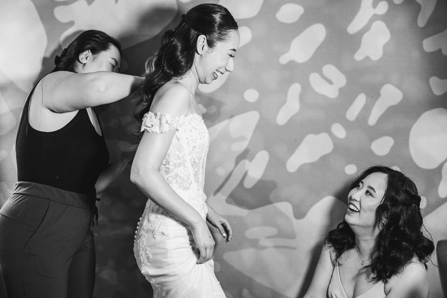 Wedding preparation moment with a bride and helpers against a patterned light backdrop.