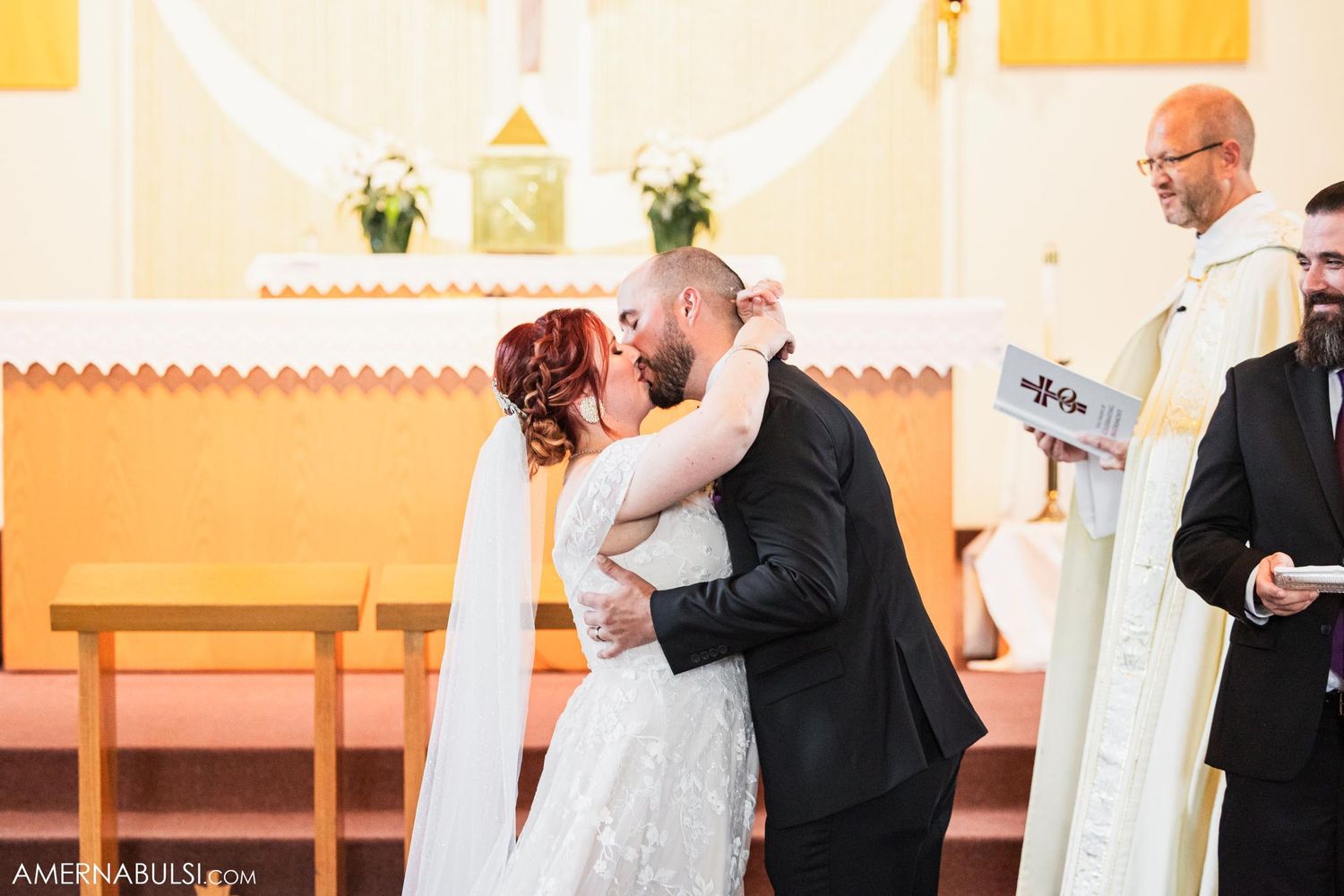 Wedding couple sharing their first kiss as newlyweds at altar with priest looking on.